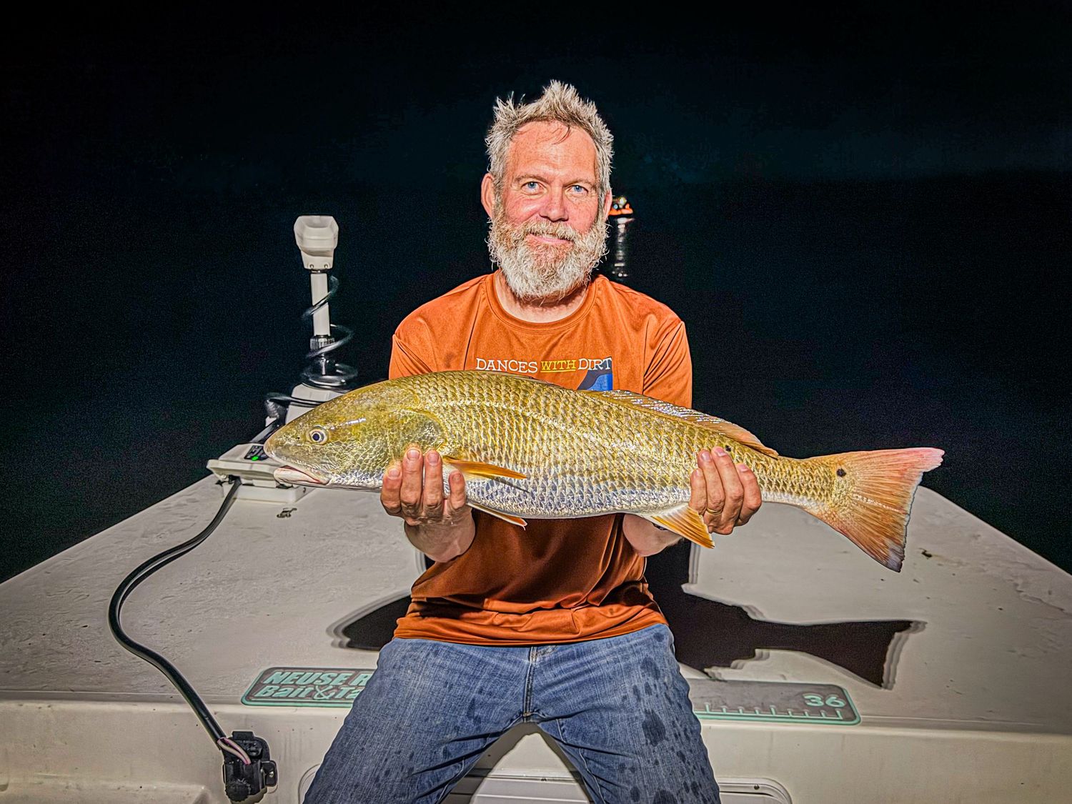 Man with white beard in orange shirt proudly holds a large redfish on a boat at night.