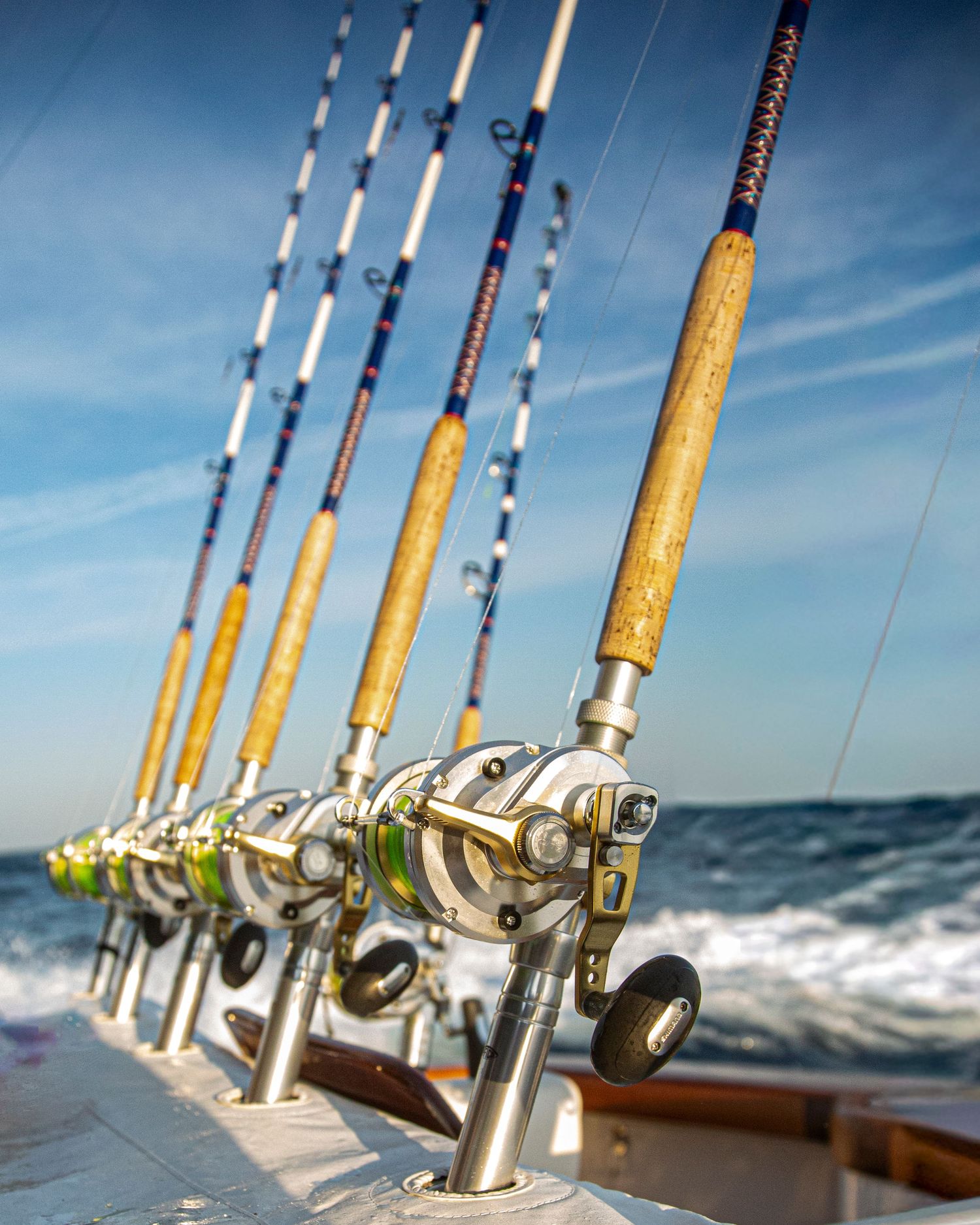 A row of deep-sea fishing rods with chrome reels lined up on a boat rail with ocean waves in the background.