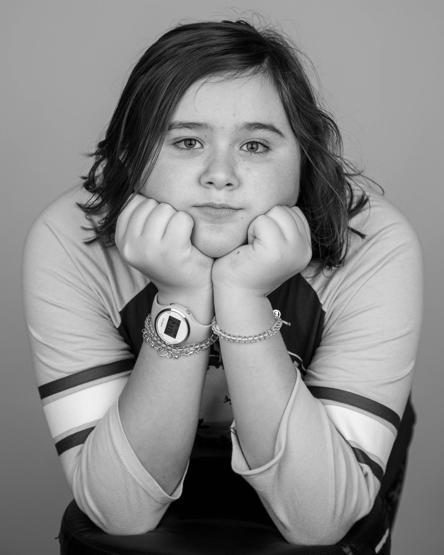 Black and white portrait of a young girl resting chin on hands, wearing a watch and bracelet, smiling softly.