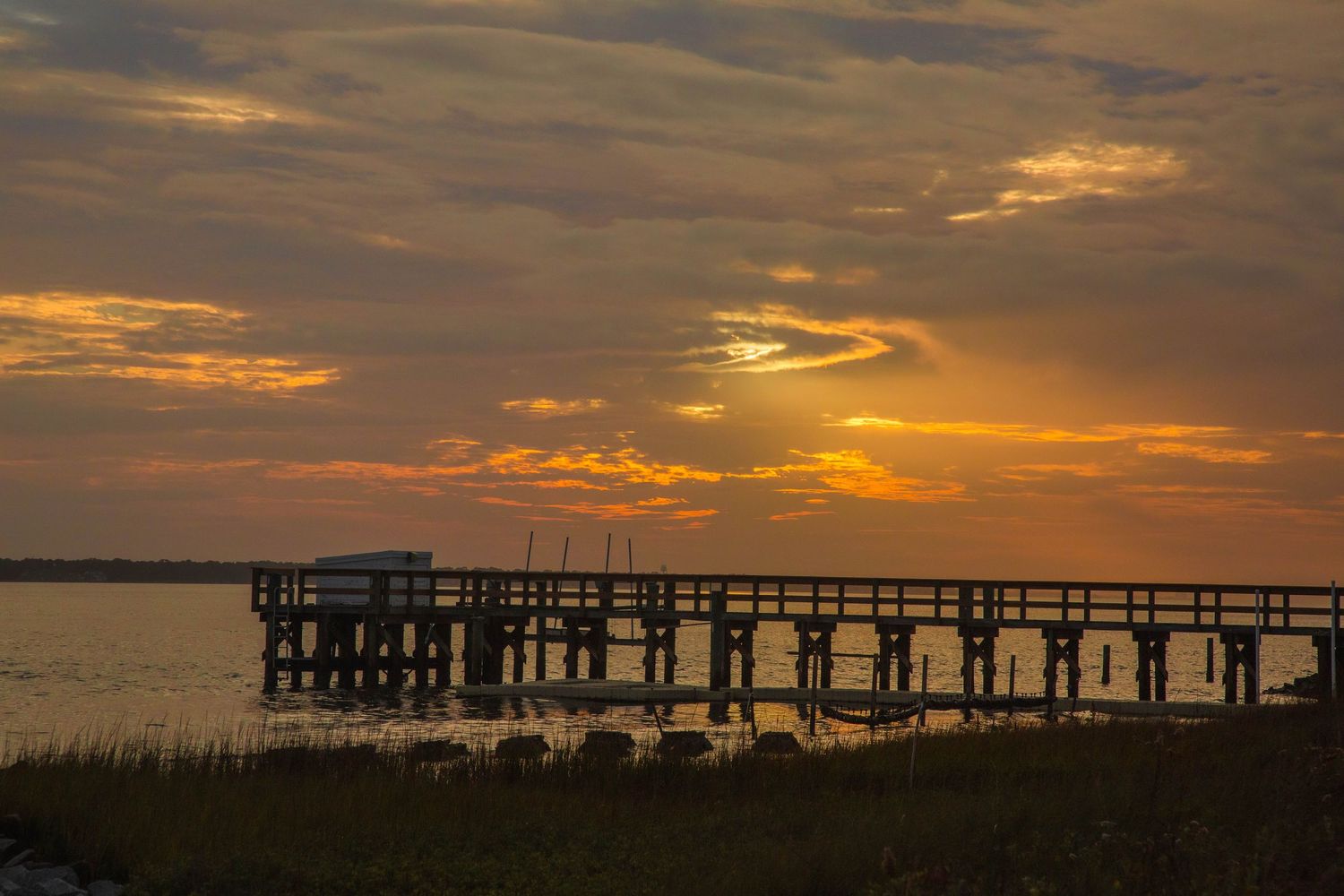 Long wooden pier silhouetted against vibrant orange sunset sky over the water.