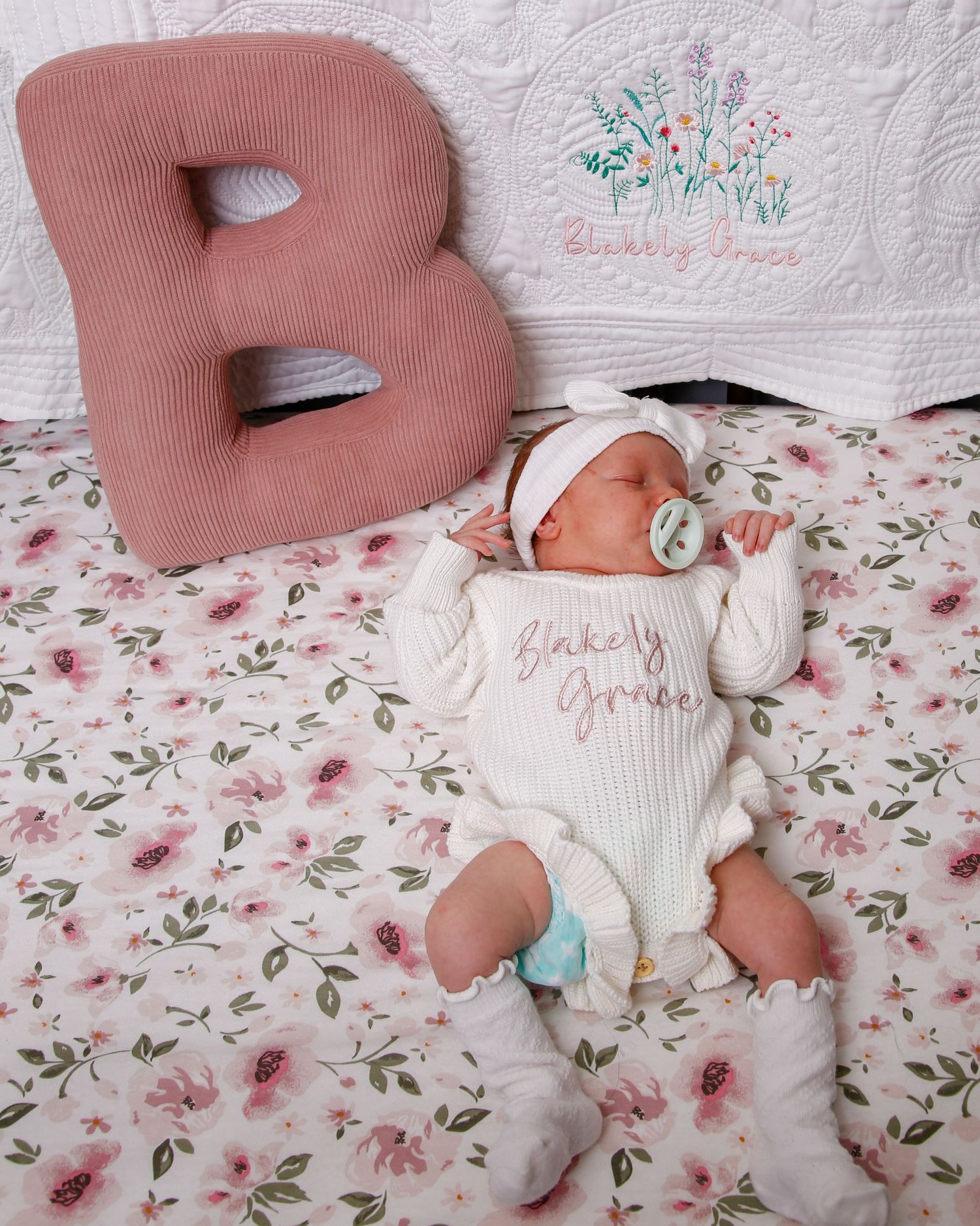 Baby sleeping on floral bedding next to large pink letter B cushion with embroidered decor in background.