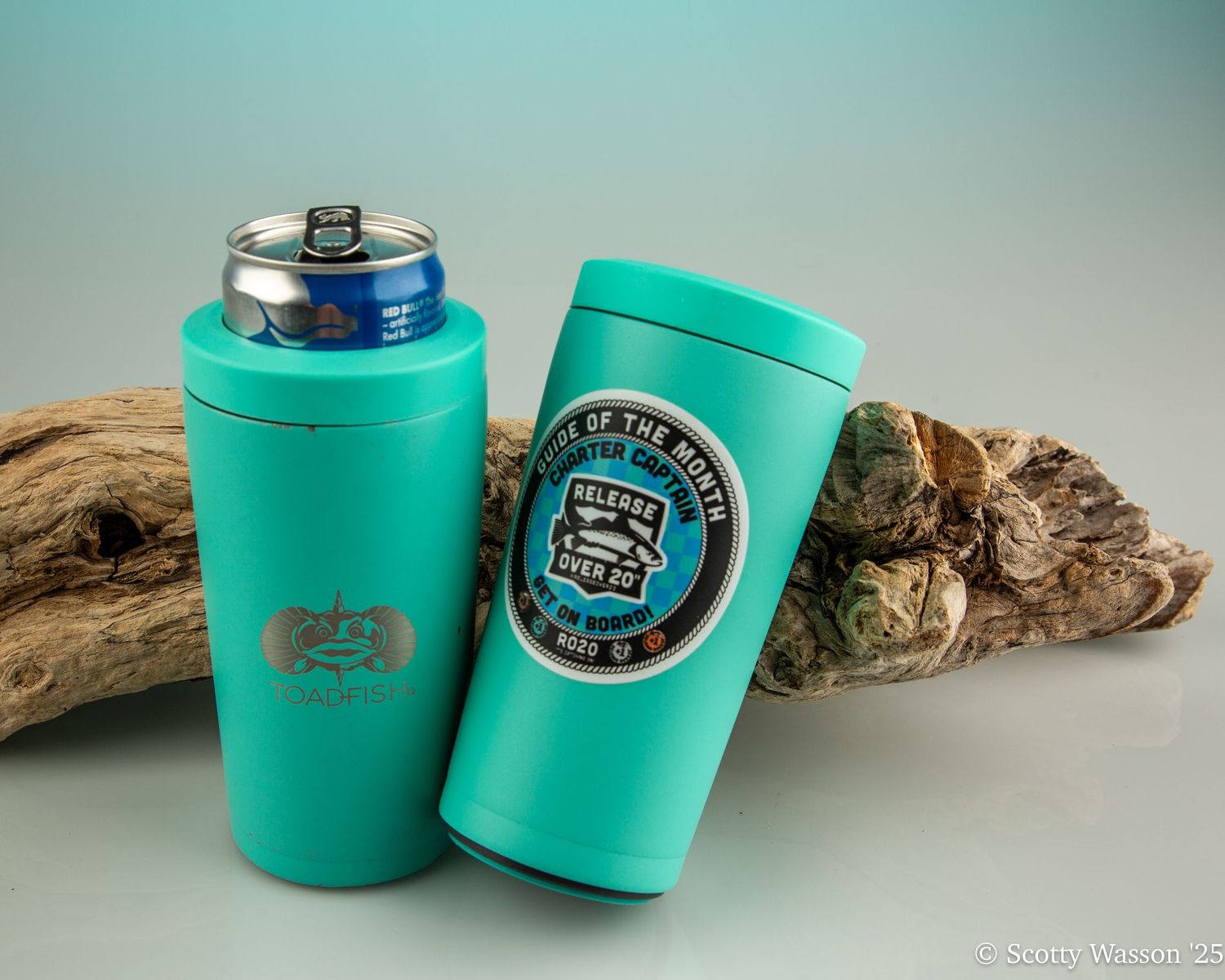 Two teal insulated can coolers with decorative patches displayed on driftwood against a light blue background.