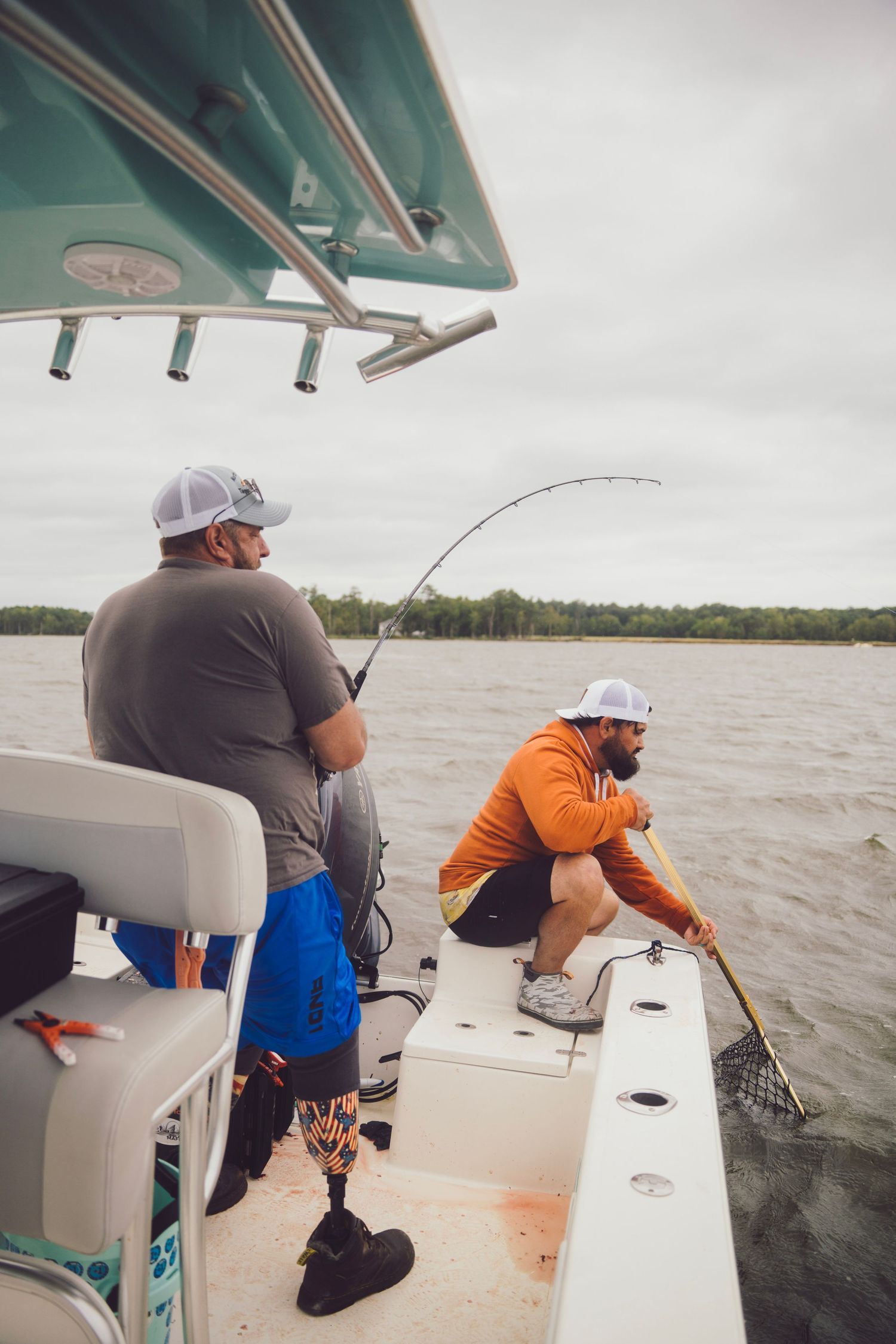 Two men fishing from a boat on calm water, one with a prosthetic leg holding a rod, the other using a landing net.