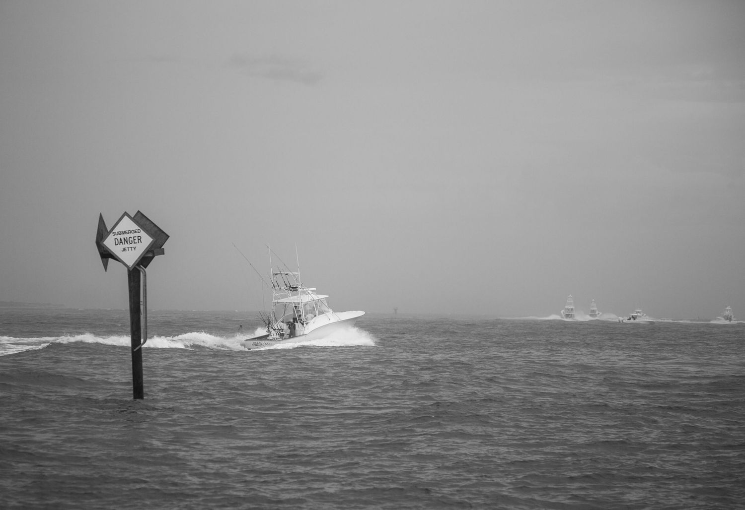 Black and white photo of fishing boats navigating past channel markers in stormy seas.
