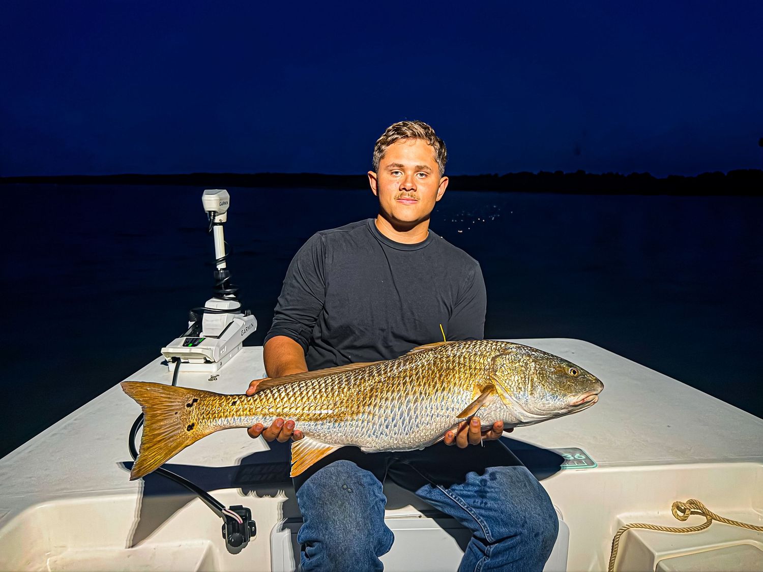 Man holding a large redfish on a boat at night with dark blue sky in the background.