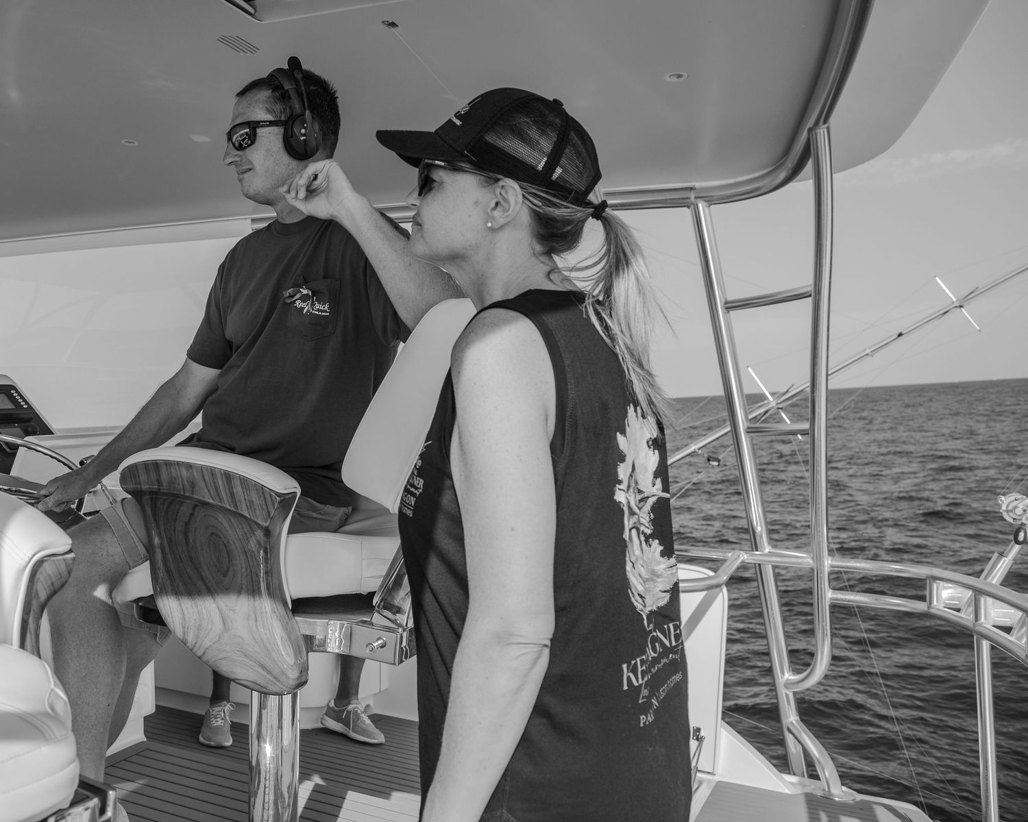 Man and woman in matching black shirts navigating a boat on open water, wearing caps on a sunny day.