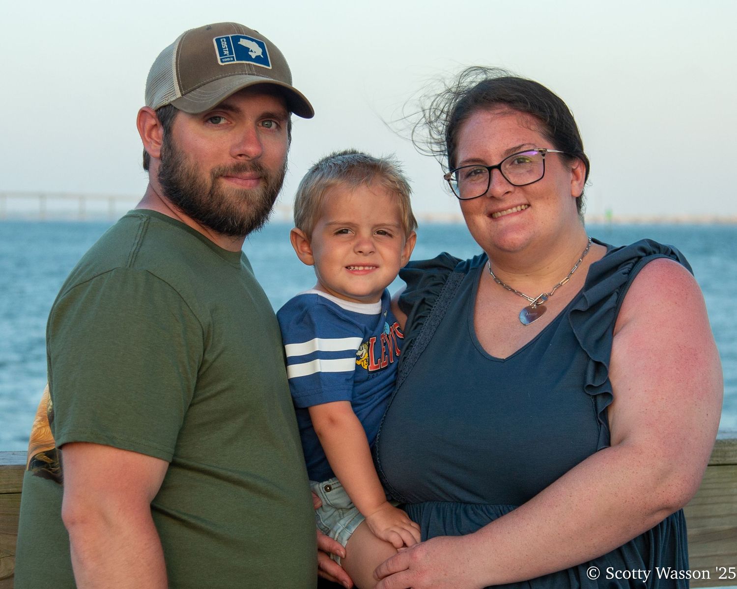 A family of three poses on a waterfront dock at sunset, with parents holding a young toddler boy between them.