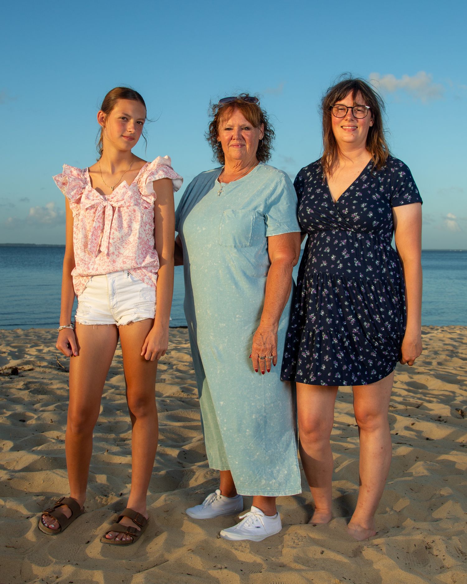 Three women of different ages pose together on a sandy beach at sunset, wearing casual summer outfits.