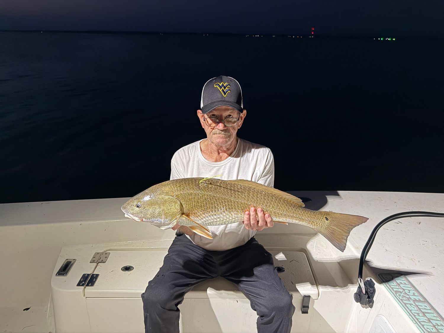 Angler on a boat at night proudly holds a large redfish caught during a nighttime fishing trip on open water.