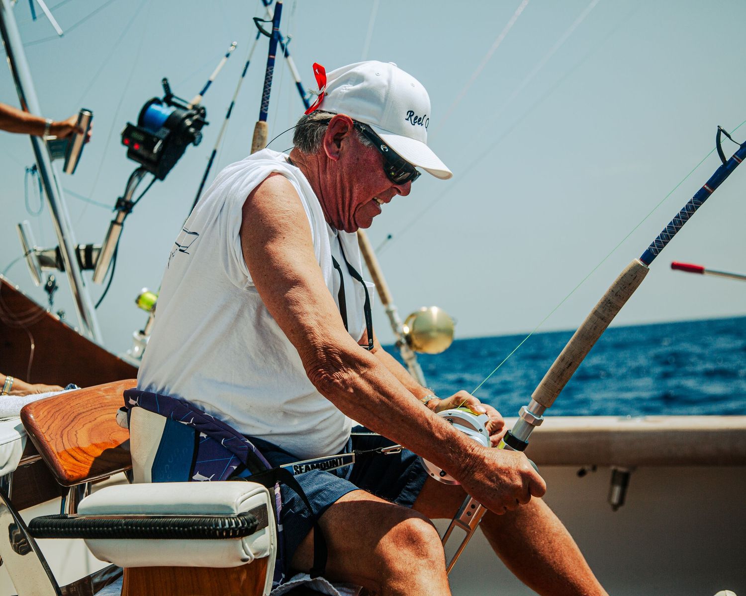 Man in white cap and sleeveless shirt fishing on a boat in calm blue ocean waters on a sunny day.
