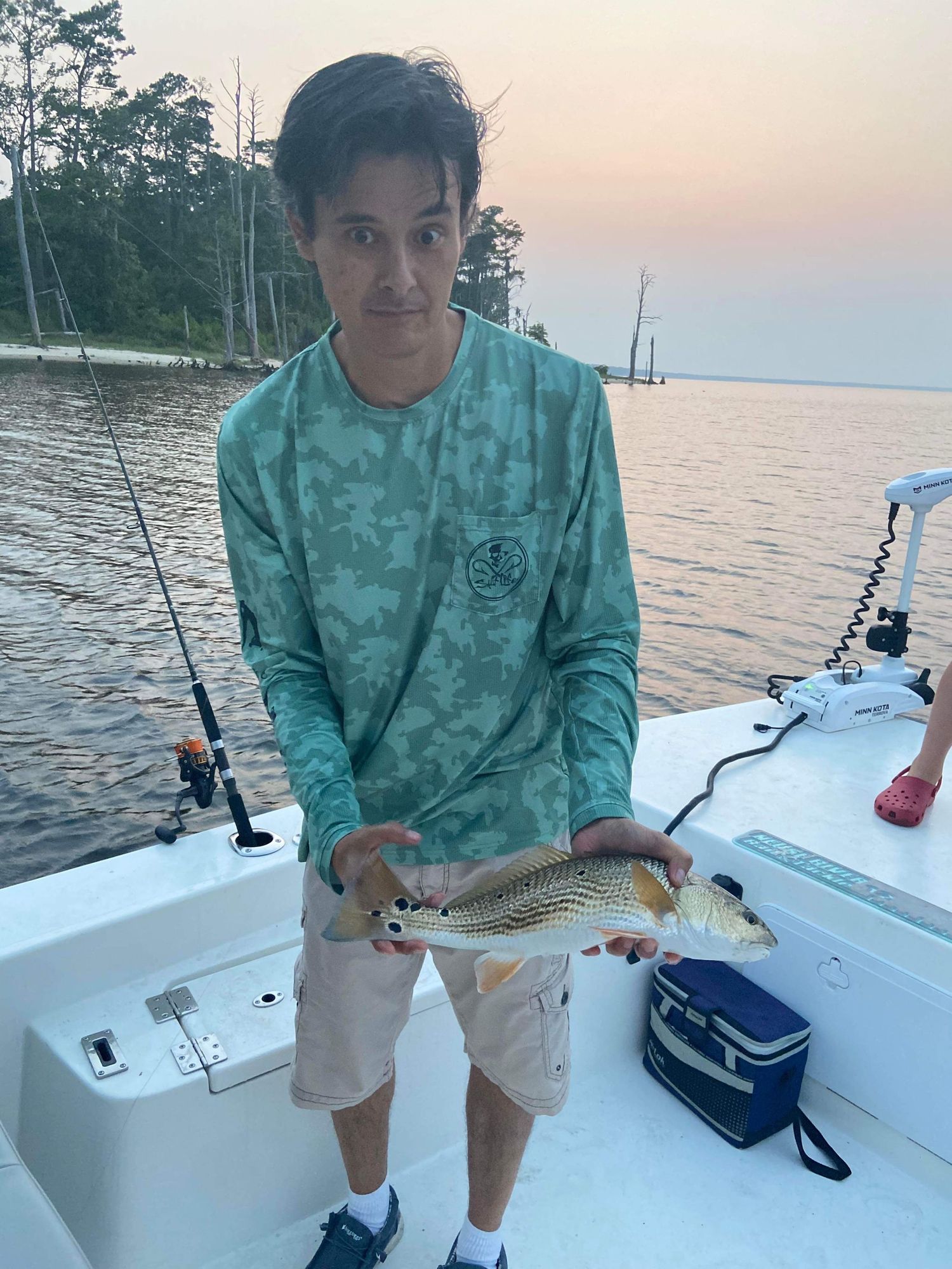 Angler in teal camo shirt holds up a freshly caught fish on a boat during sunset.