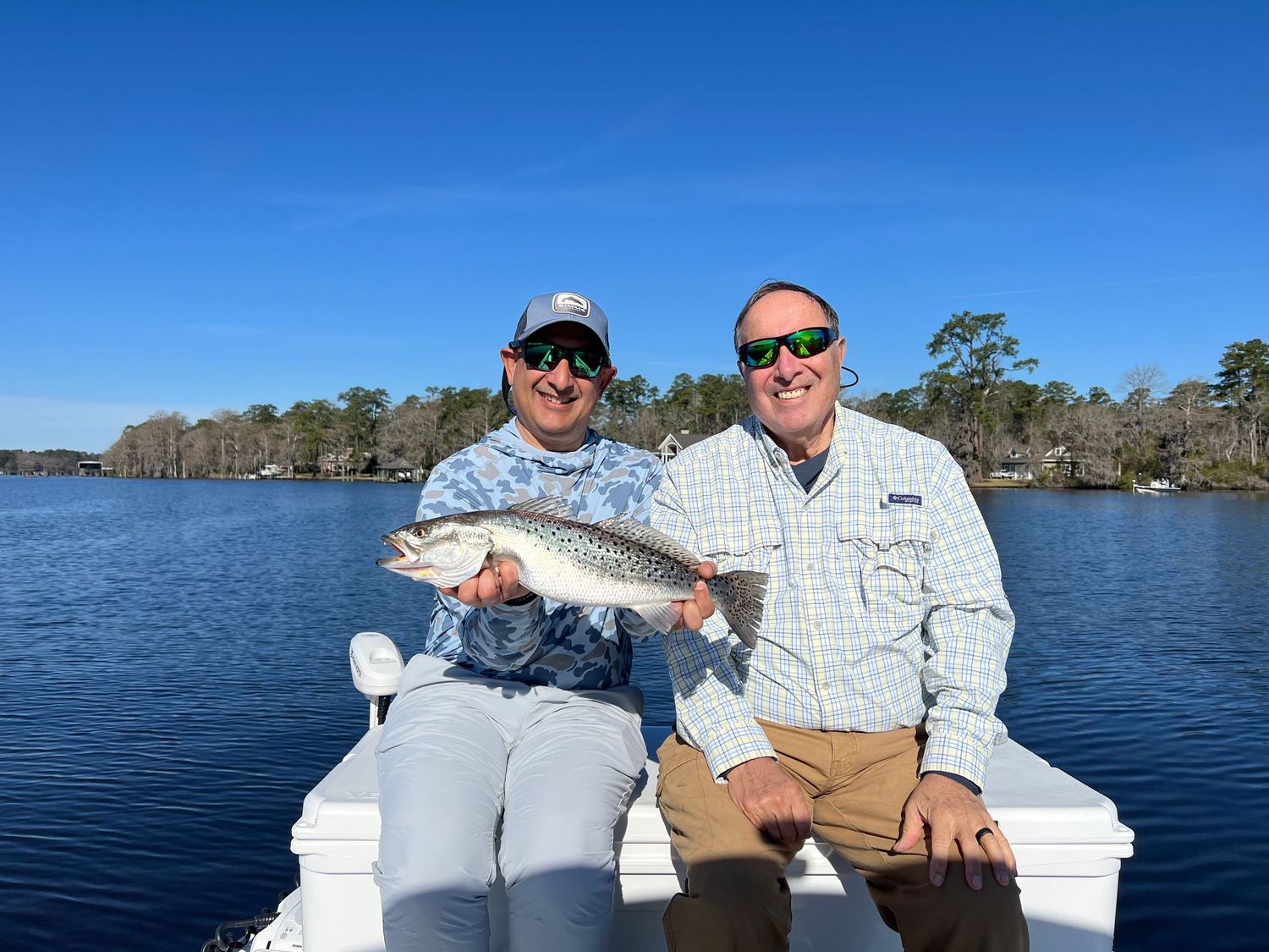 Two anglers pose with caught fish on white boat deck with coastal waters and trees behind them.