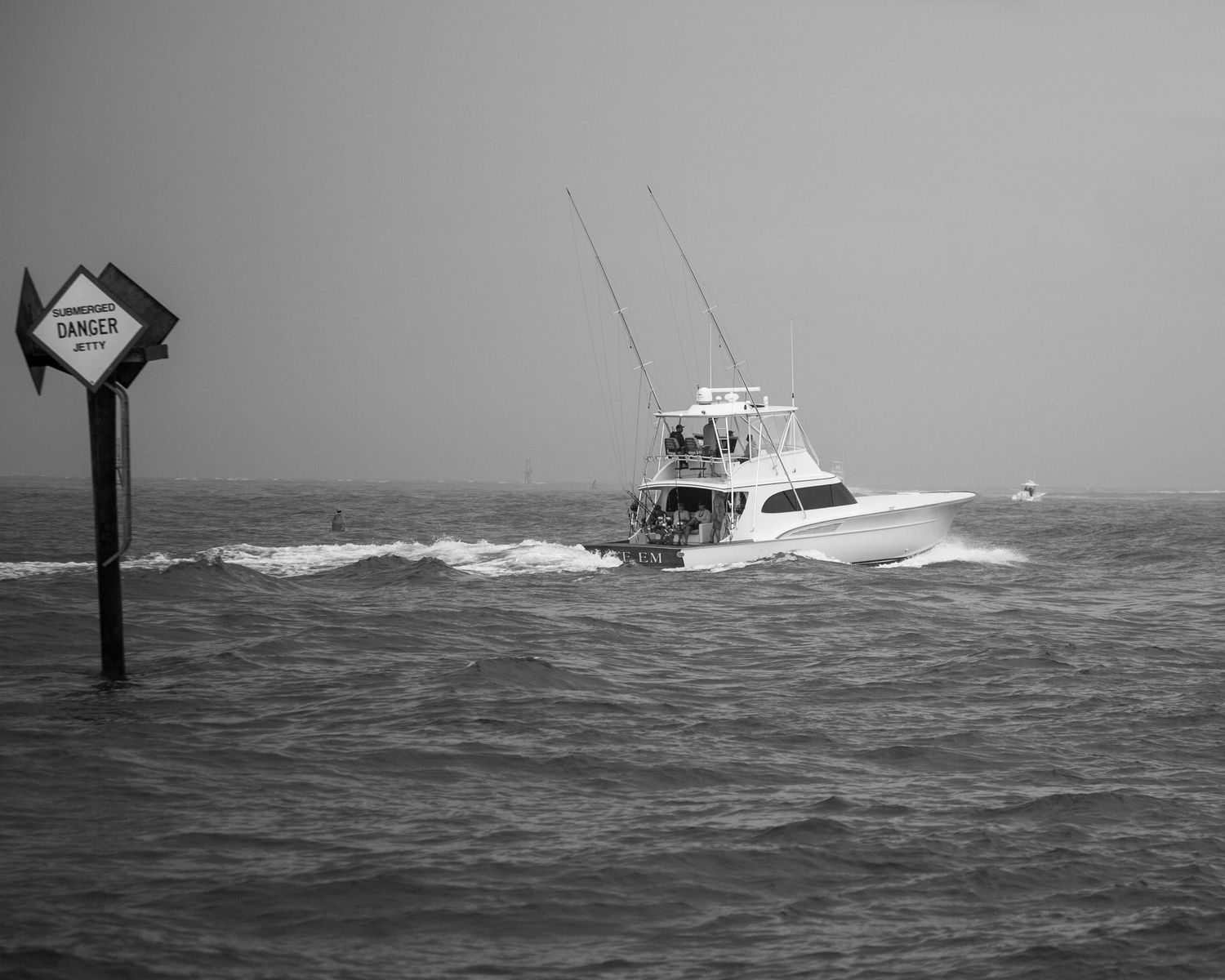 Black and white image of a fishing boat navigating rough waters past a warning sign.