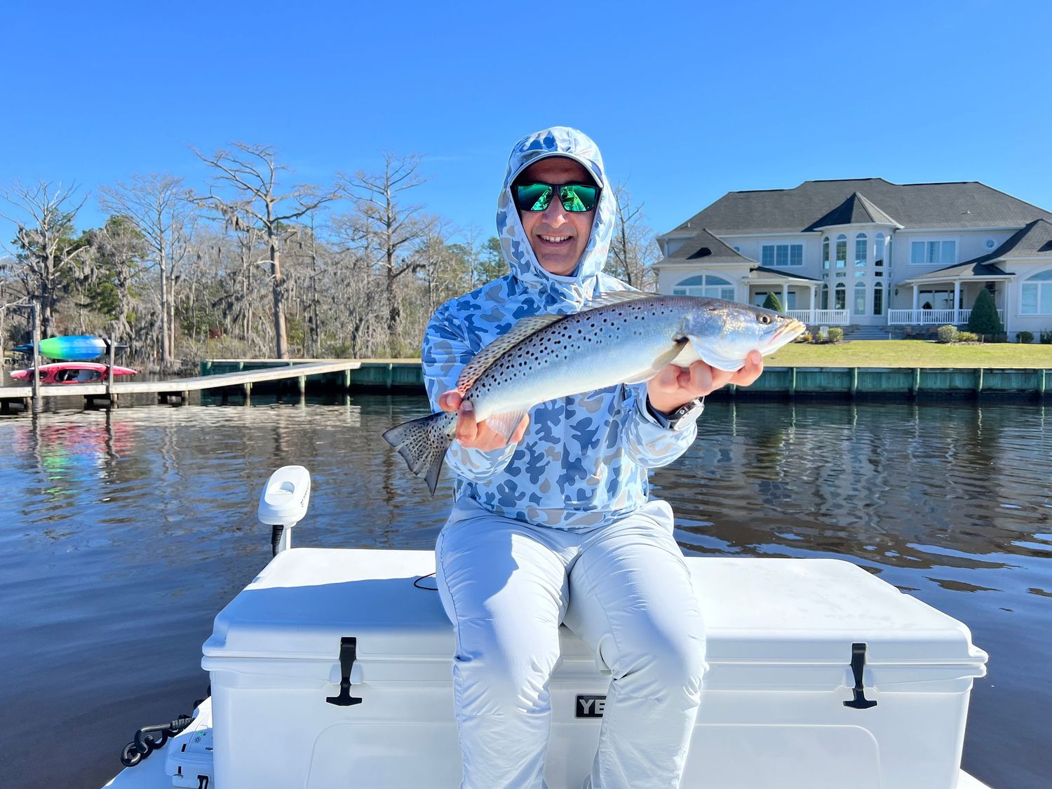 Angler in blue camouflage shirt displays red drum catch while sitting on cooler by waterfront house.
