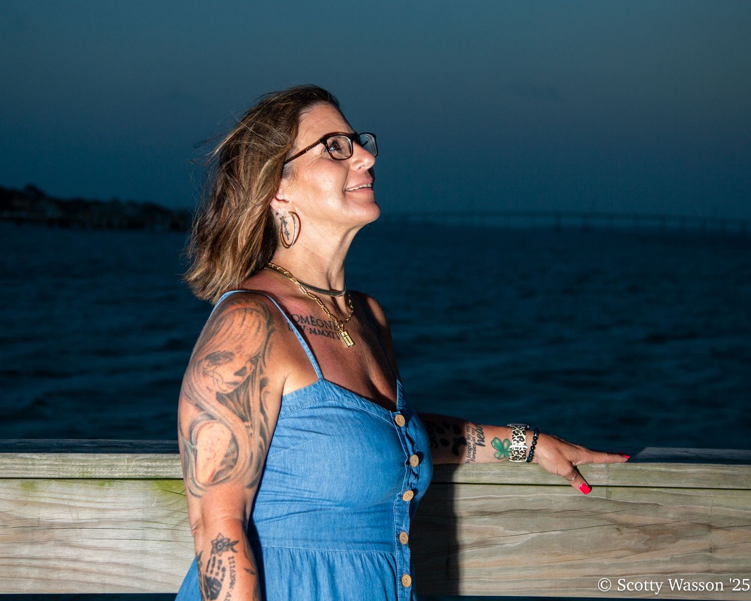 Woman with tattoos and glasses in blue dress smiling at twilight by the ocean.