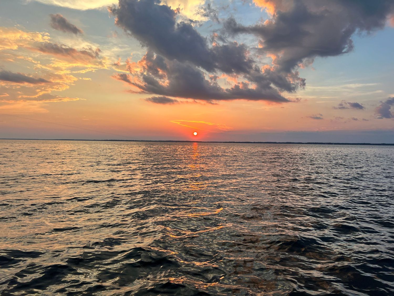 Vibrant orange sun sets over dark rippling ocean waters beneath dramatic cloudy sky at dusk.