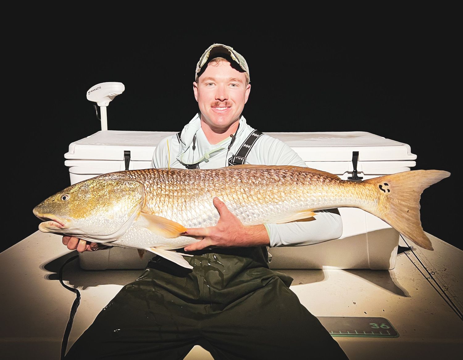 Someone holding a large redfish catch at night against a dark background with white dock lights visible.
