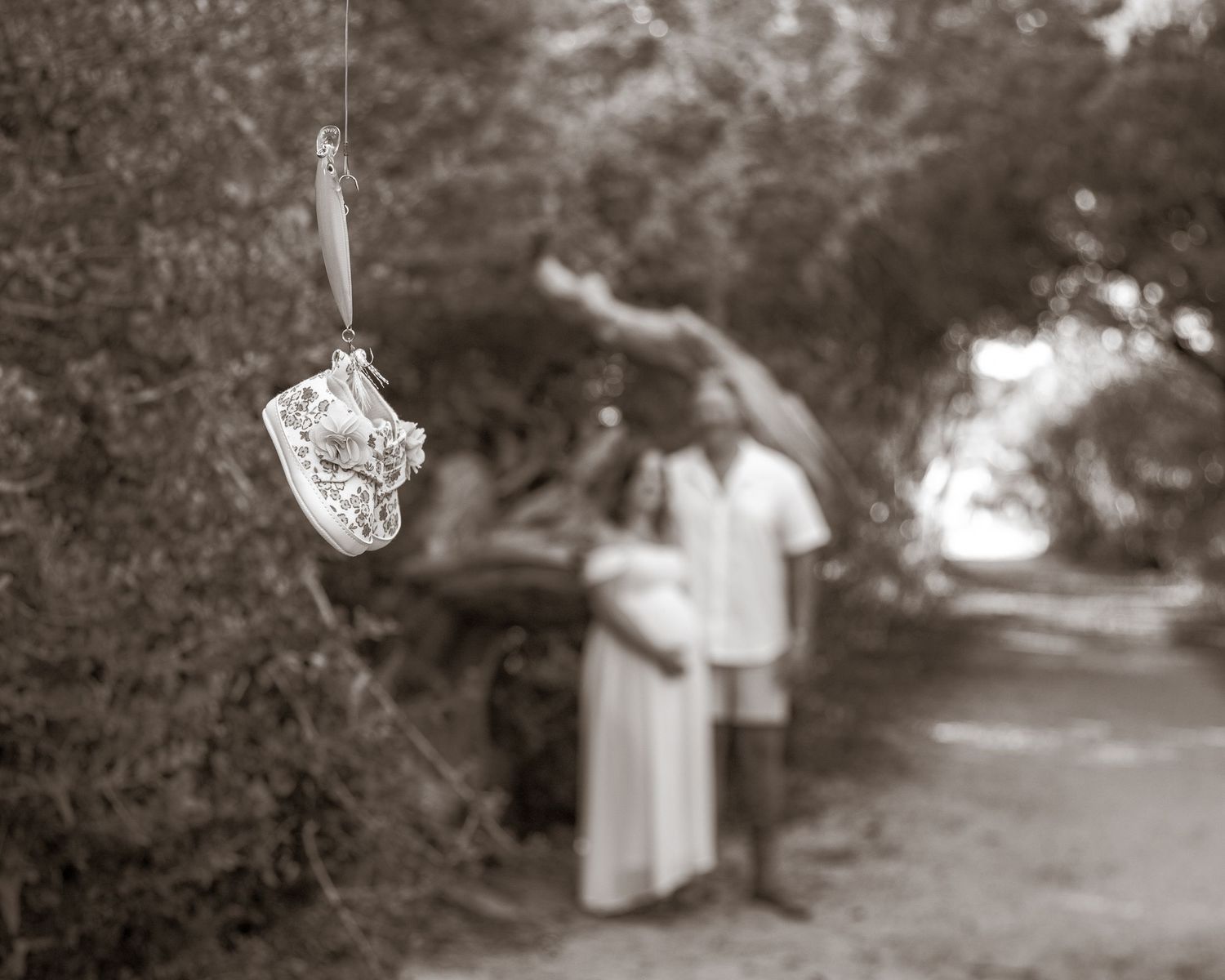 Sepia-toned maternity photo of a couple on a path, with baby shoes hanging in the foreground.