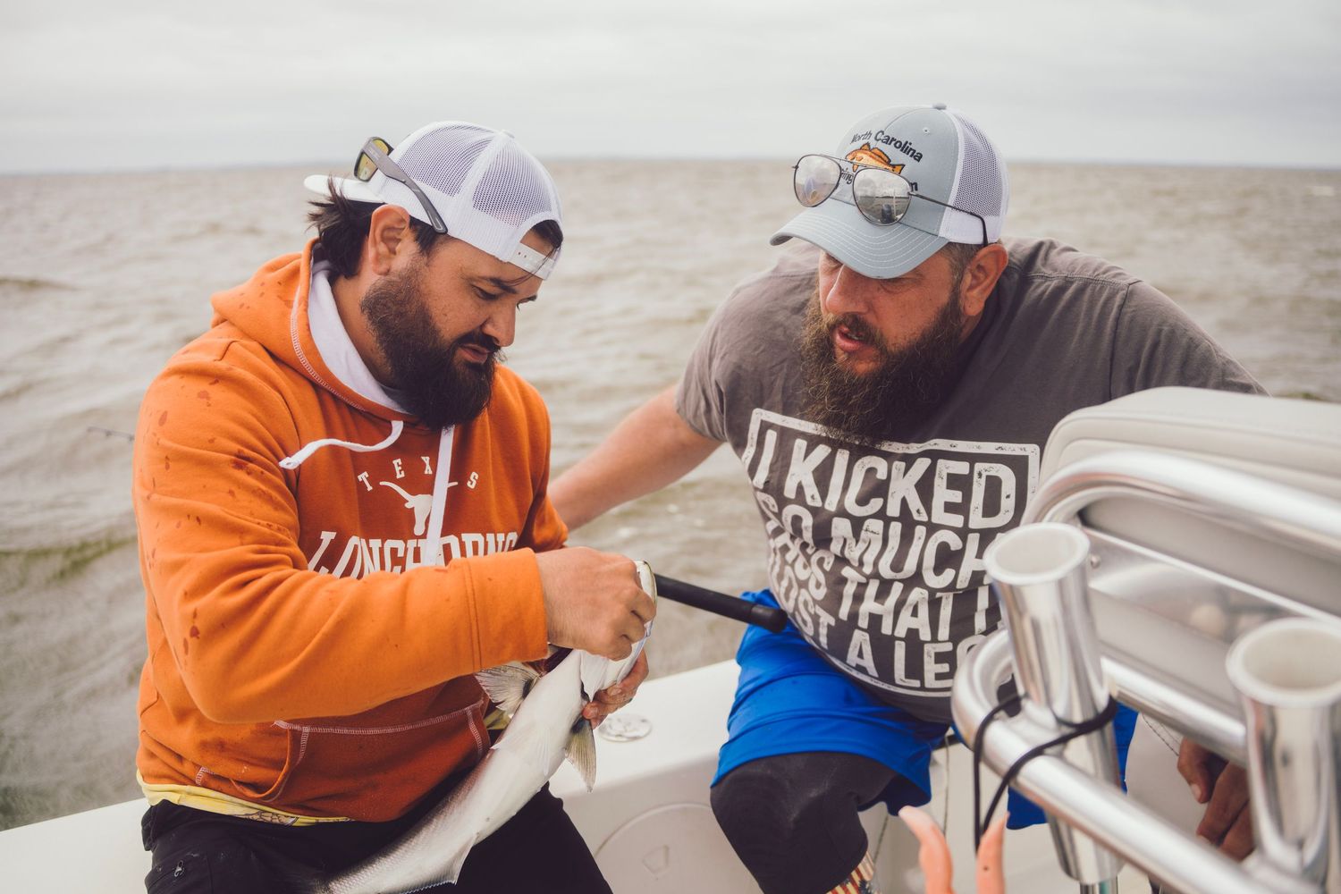 Two bearded men in trucker hats examine a fish on a boat on open water.
