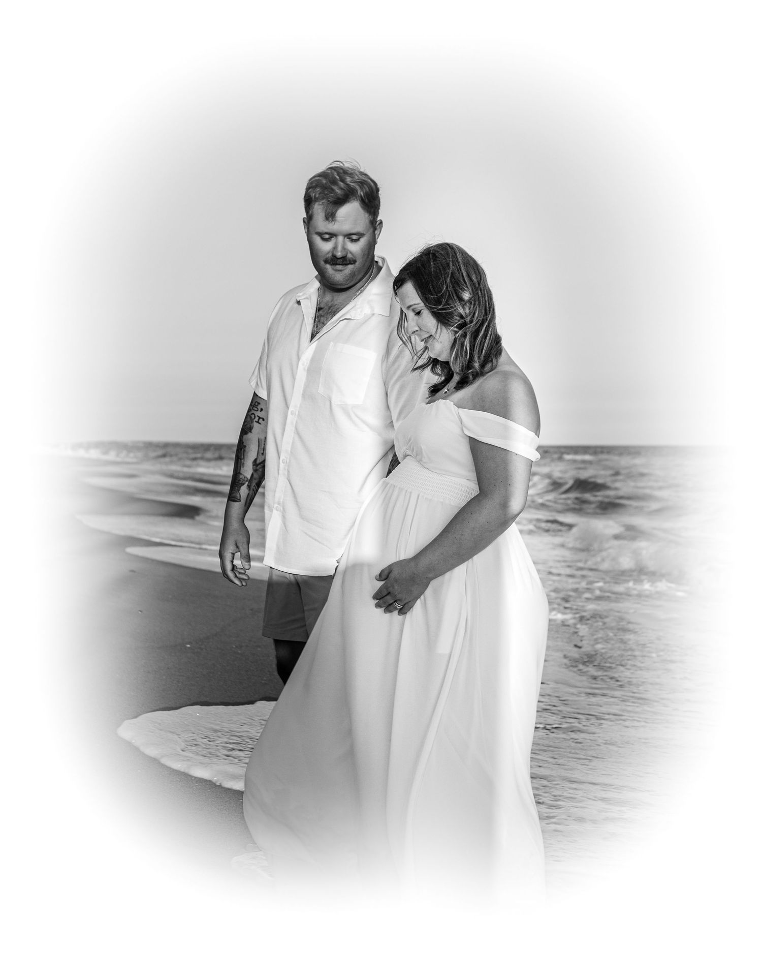 Black and white beach wedding photo of couple in white attire sharing a tender moment by the ocean waves.