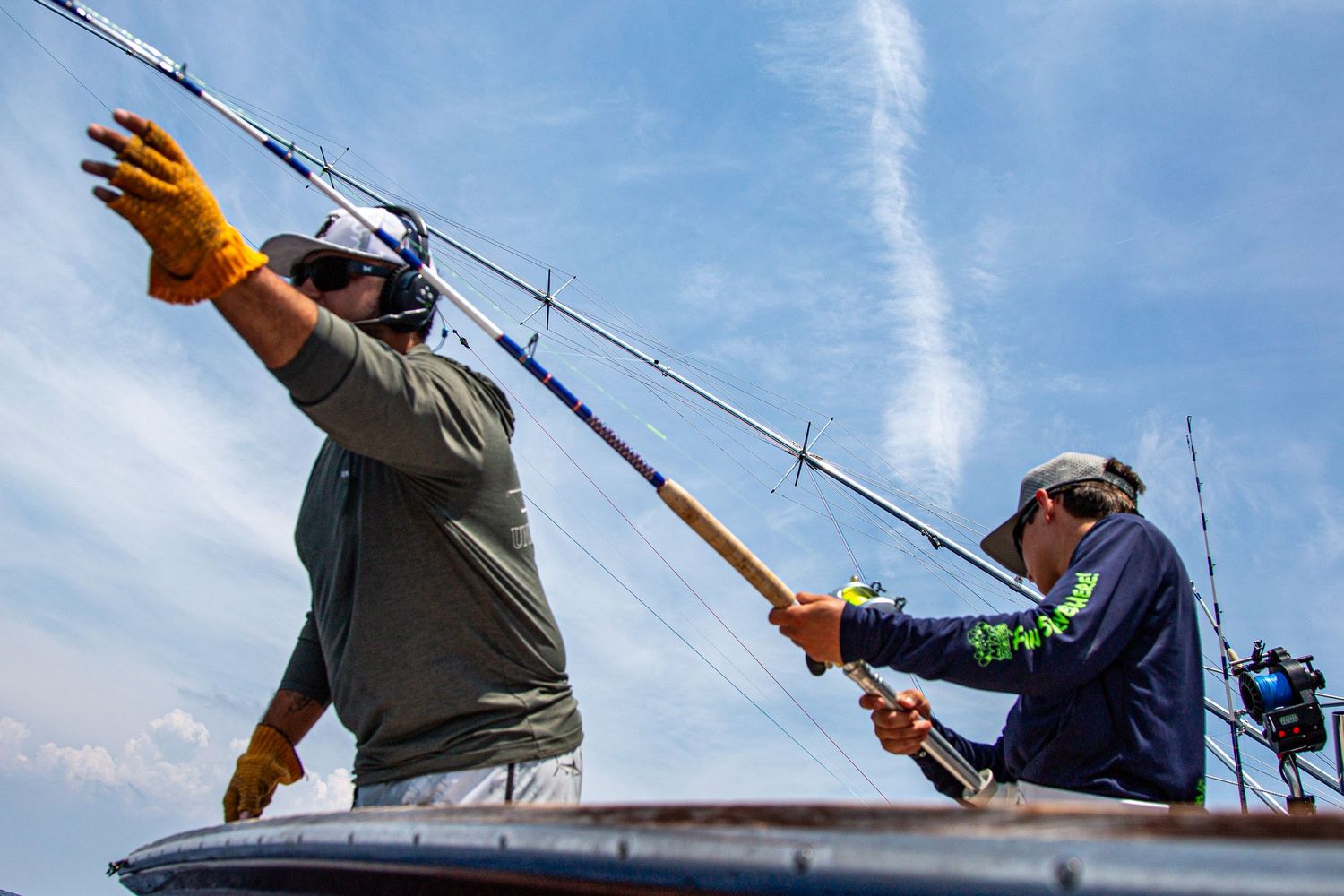 Two fishermen on a boat handling fishing rods under a blue sky, wearing caps and gloves on open water.