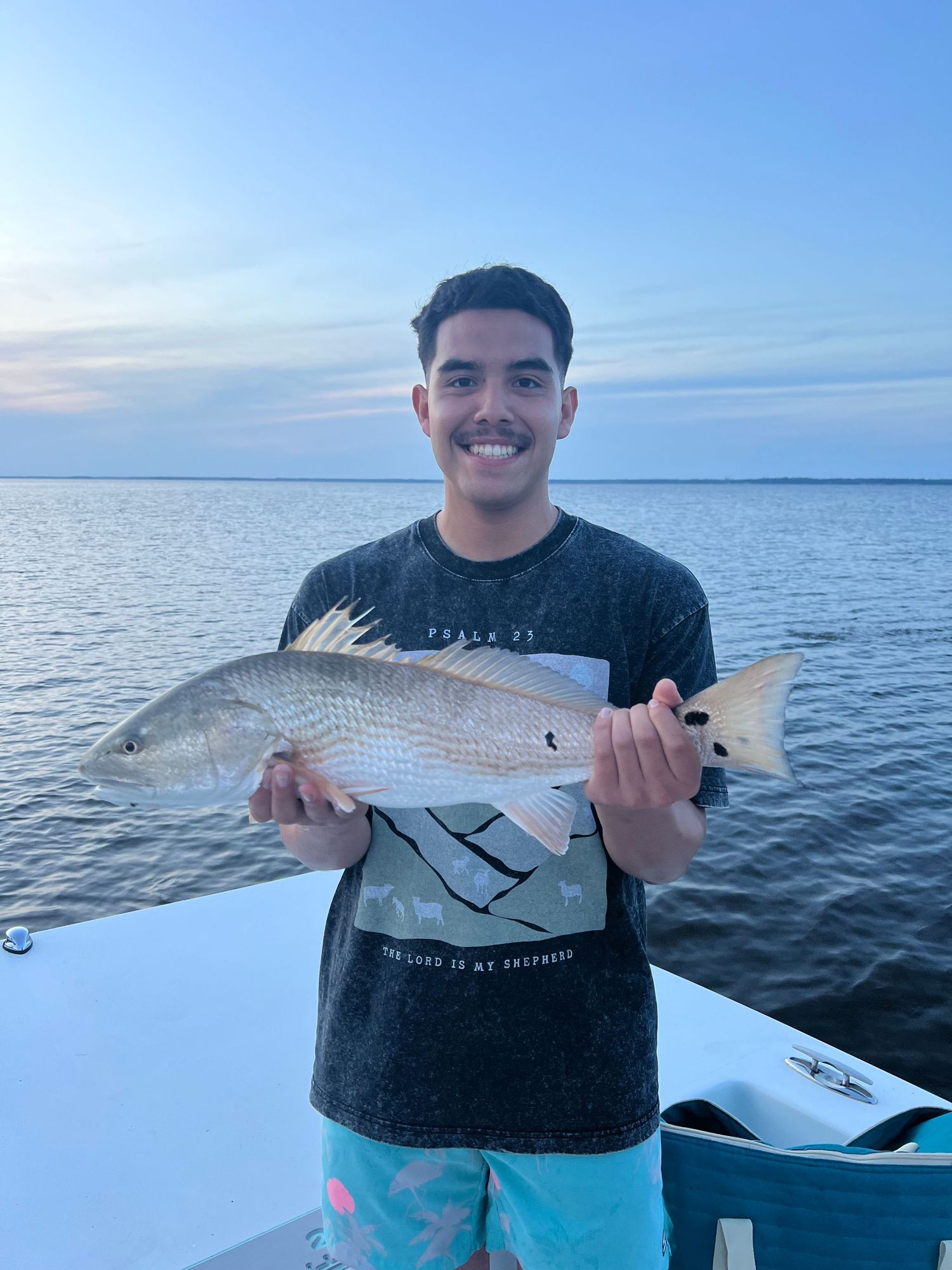 Angler on boat proudly displays large red snapper catch against calm ocean backdrop at sunset.