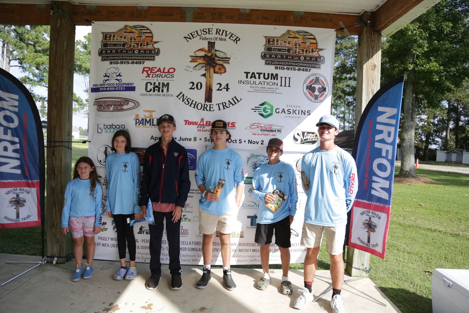 Group photo of event participants standing in front of sponsor banner at outdoor venue.