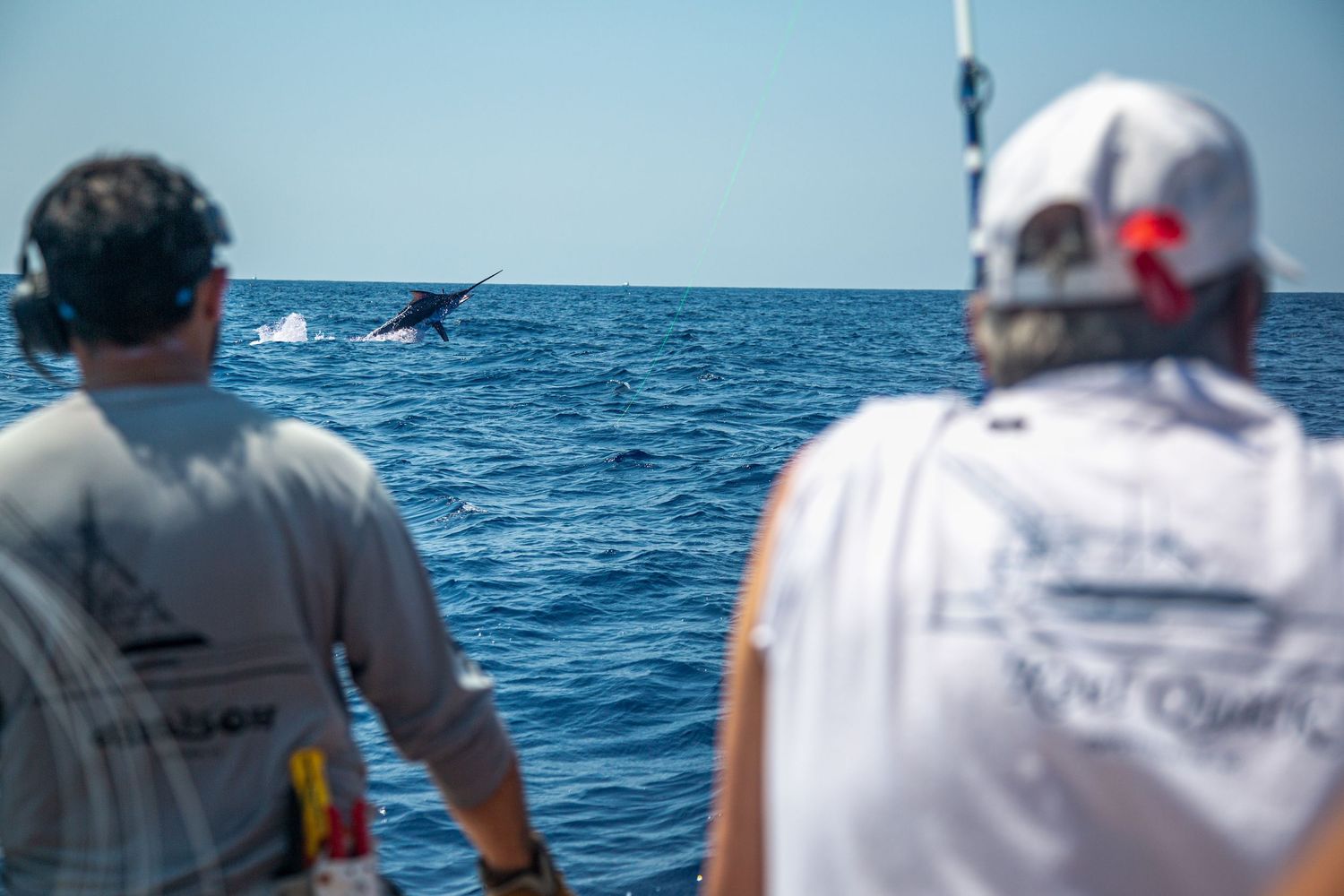 Two fishermen on a boat watching a marlin leap from the ocean on a sunny day.