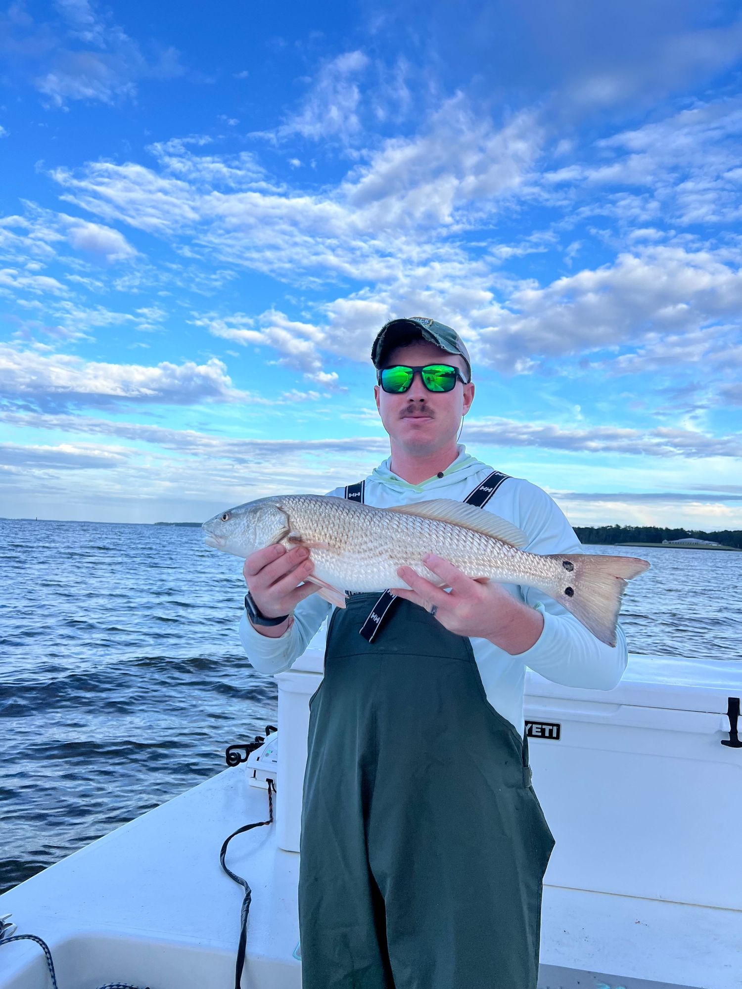 Angler in green waders and sunglasses holds freshly caught mullet fish on boat with blue sky and clouds above.
