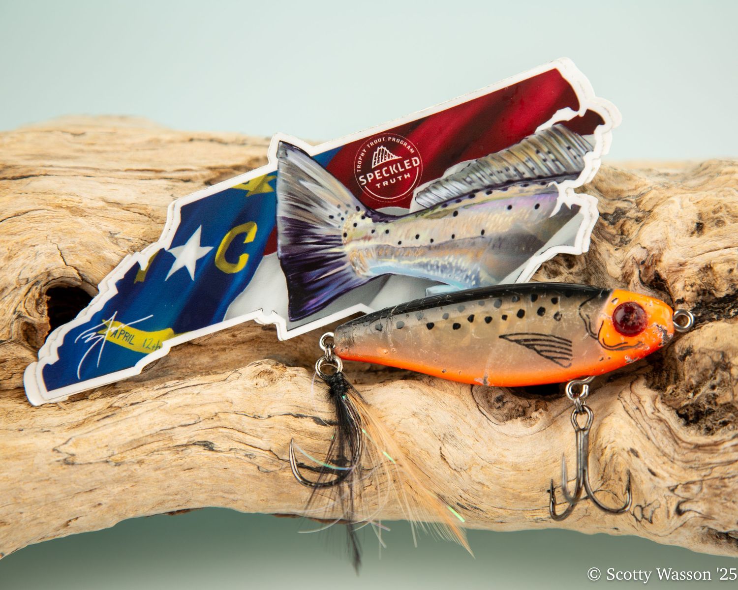 North Carolina state outline sticker with speckled trout design next to an orange fishing lure on driftwood.