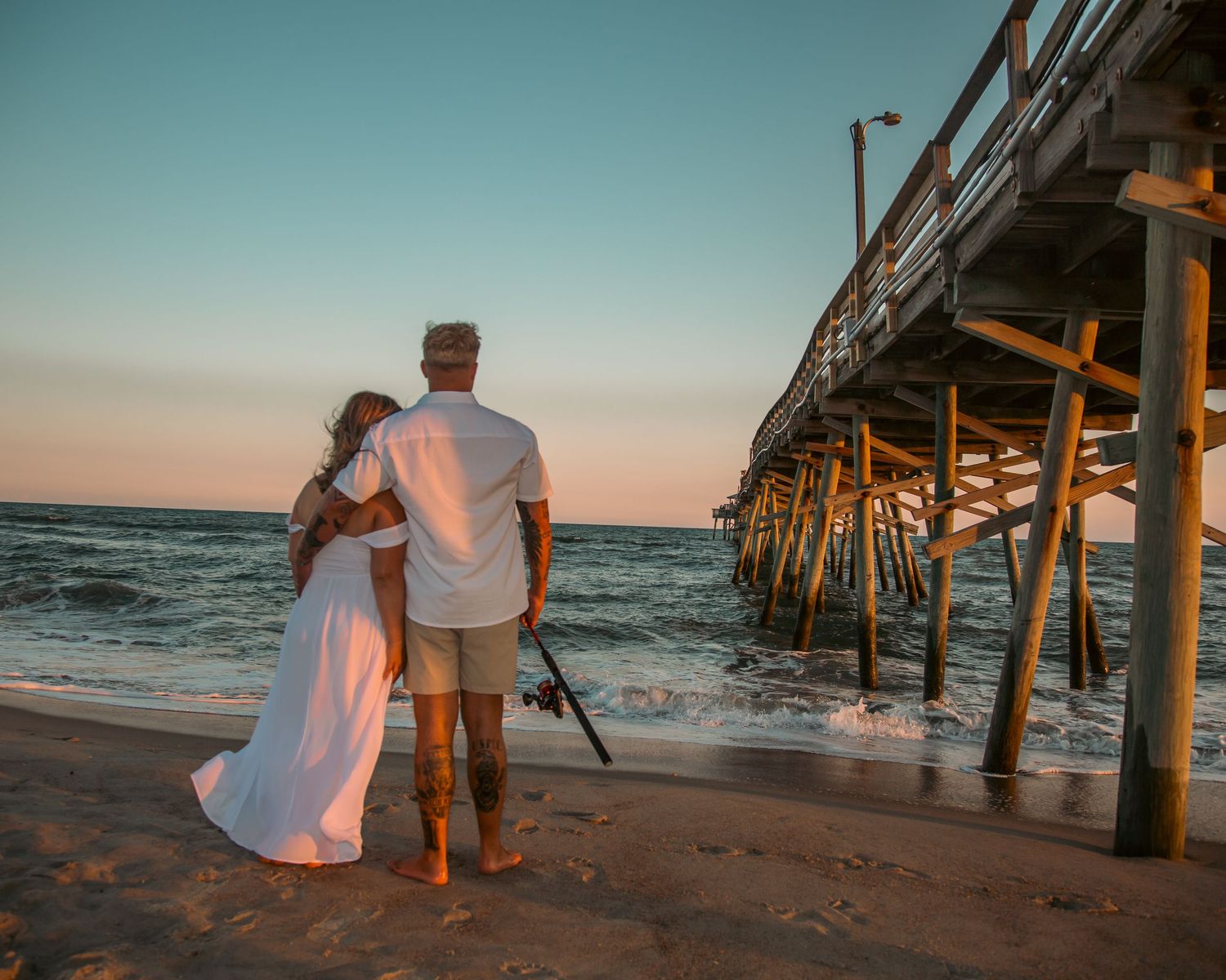 Couple in white embracing at sunset by a wooden beach pier, with ocean waves and golden sky.