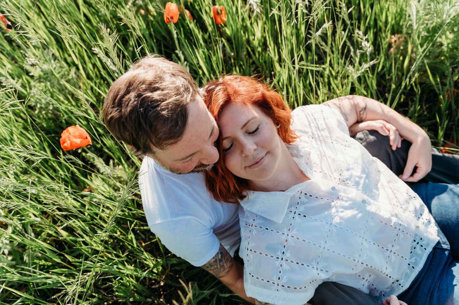 A couple relaxes together in tall grass with orange poppies blooming on a sunny day.