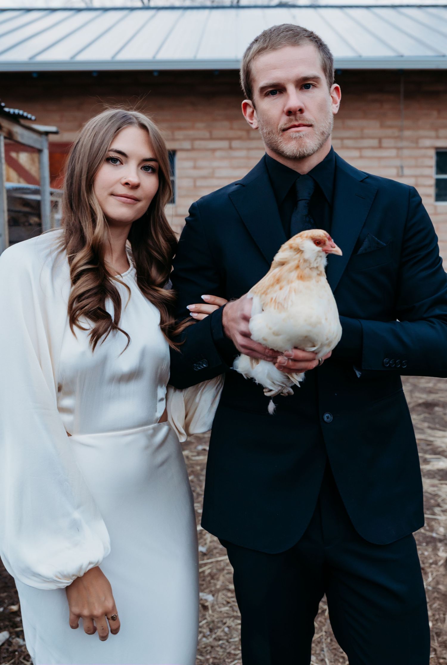 A couple in wedding attire poses outdoors while holding their white chicken pet against a brick building backdrop.
