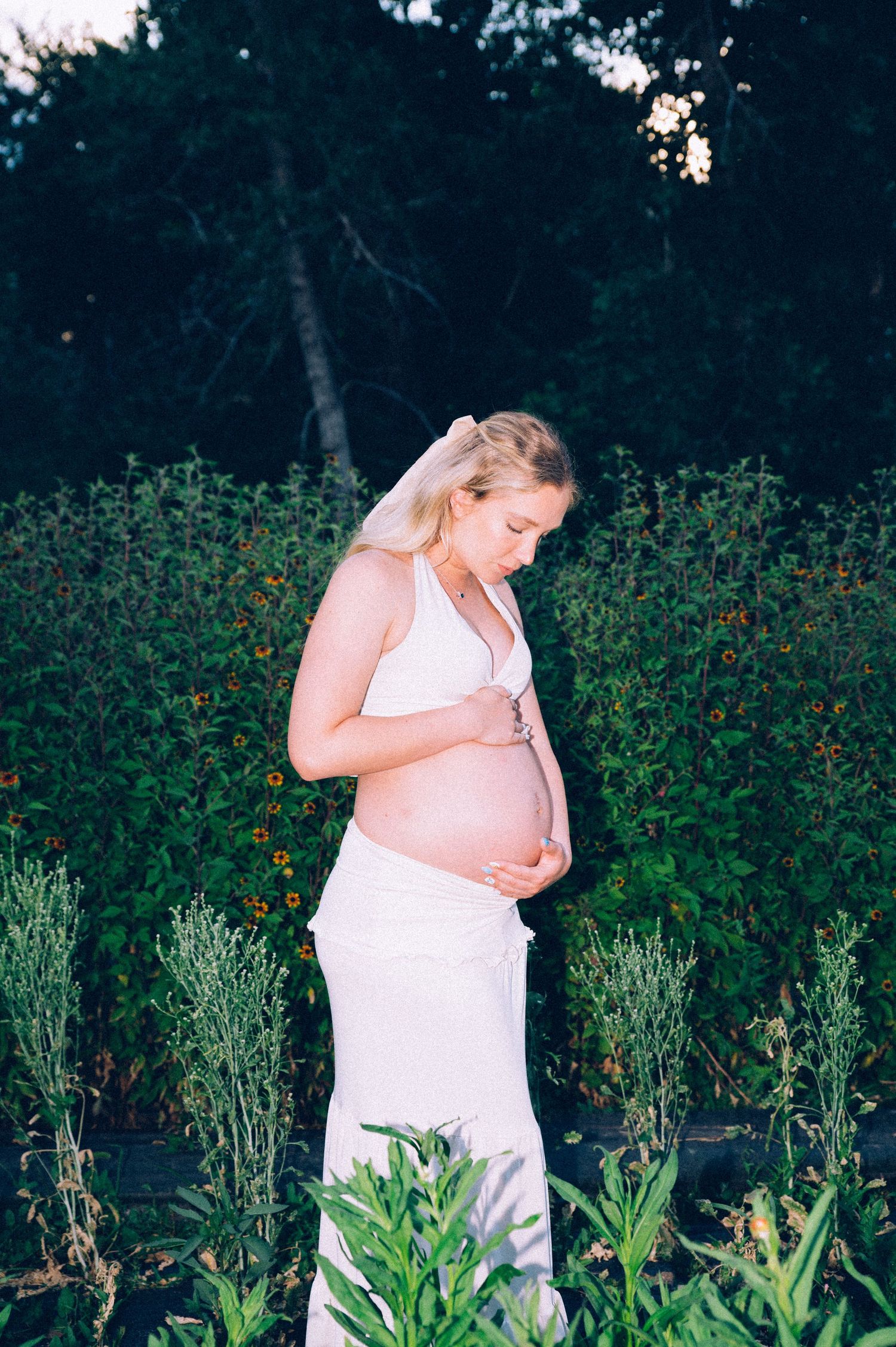 A pregnant woman in a white dress stands thoughtfully in a garden with blooming flowers at dusk.