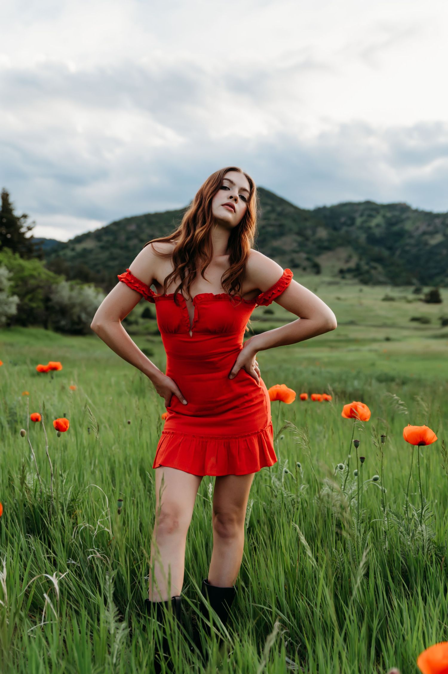 A red dress stands out against a lush green meadow with vibrant orange poppies and mountains in the background.