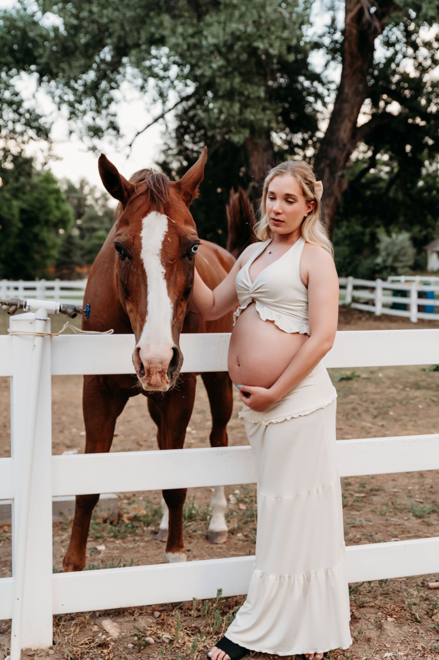 A woman in a flowing white dress poses with a brown horse by a white fence during a maternity photoshoot.