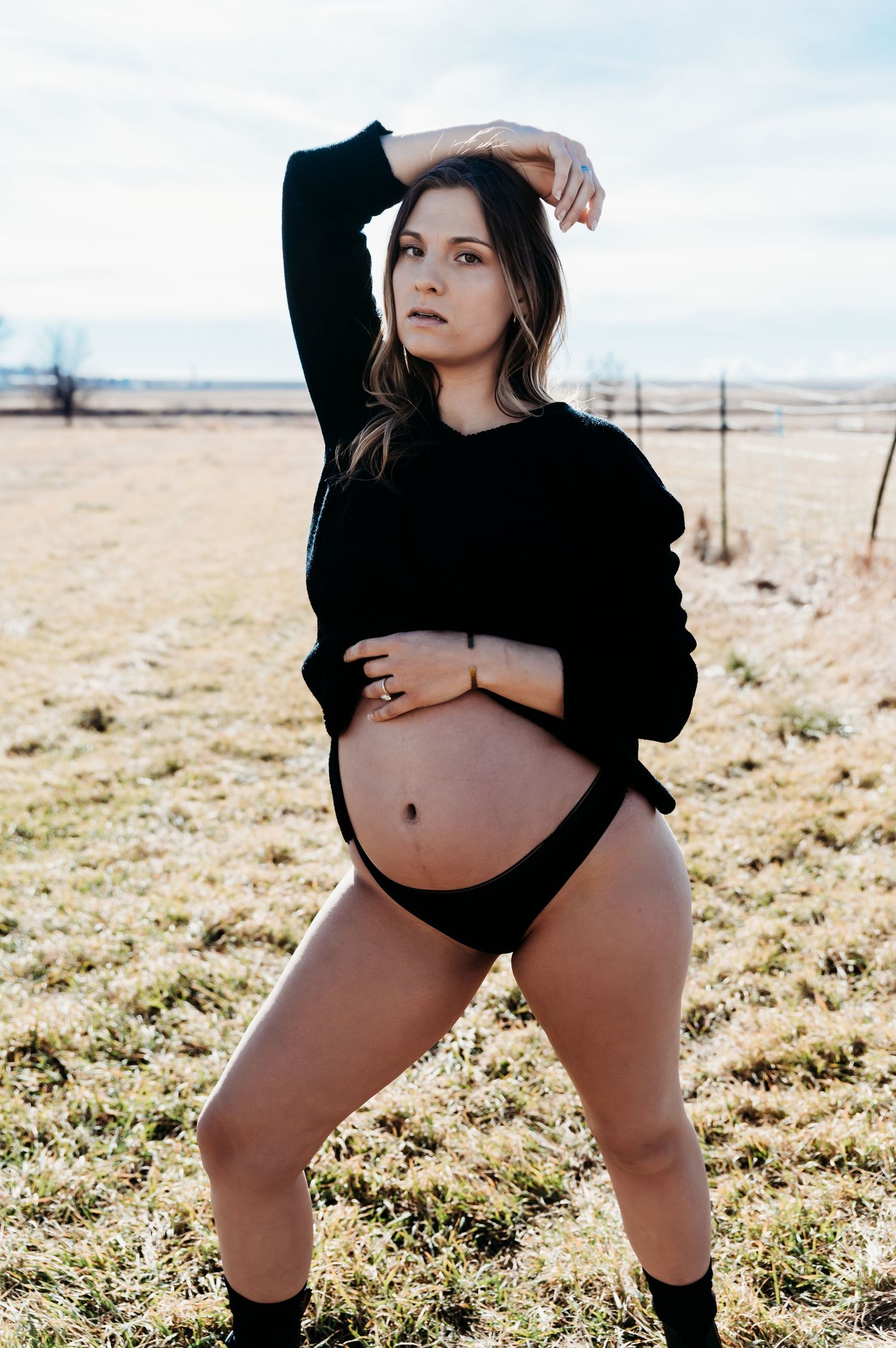 Maternity photo showing a woman in black sweater and underwear posing in an outdoor field during golden hour.