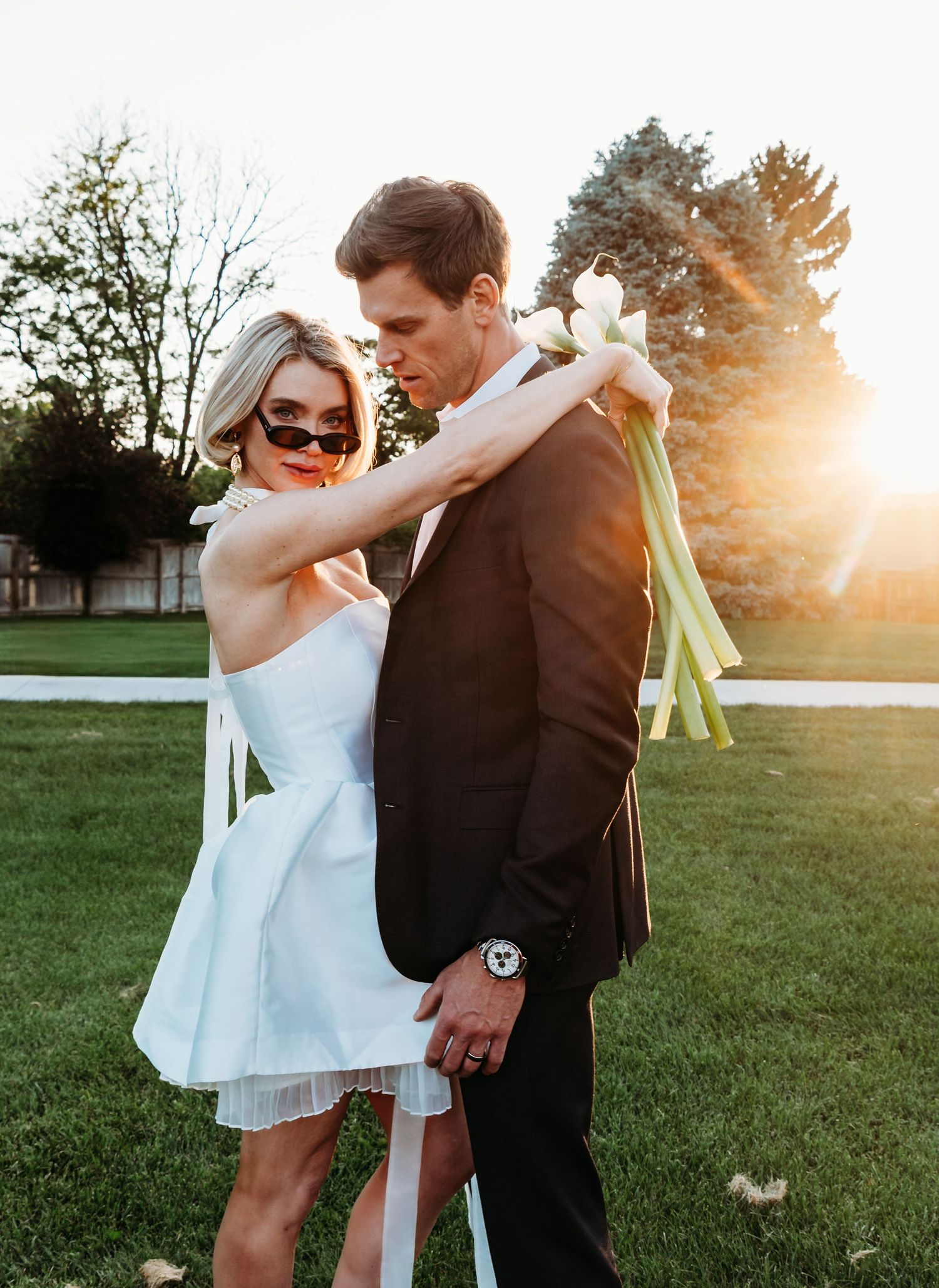 A couple embraces outdoors at sunset, with the bride wearing a short white dress and groom in black suit on a grassy lawn.