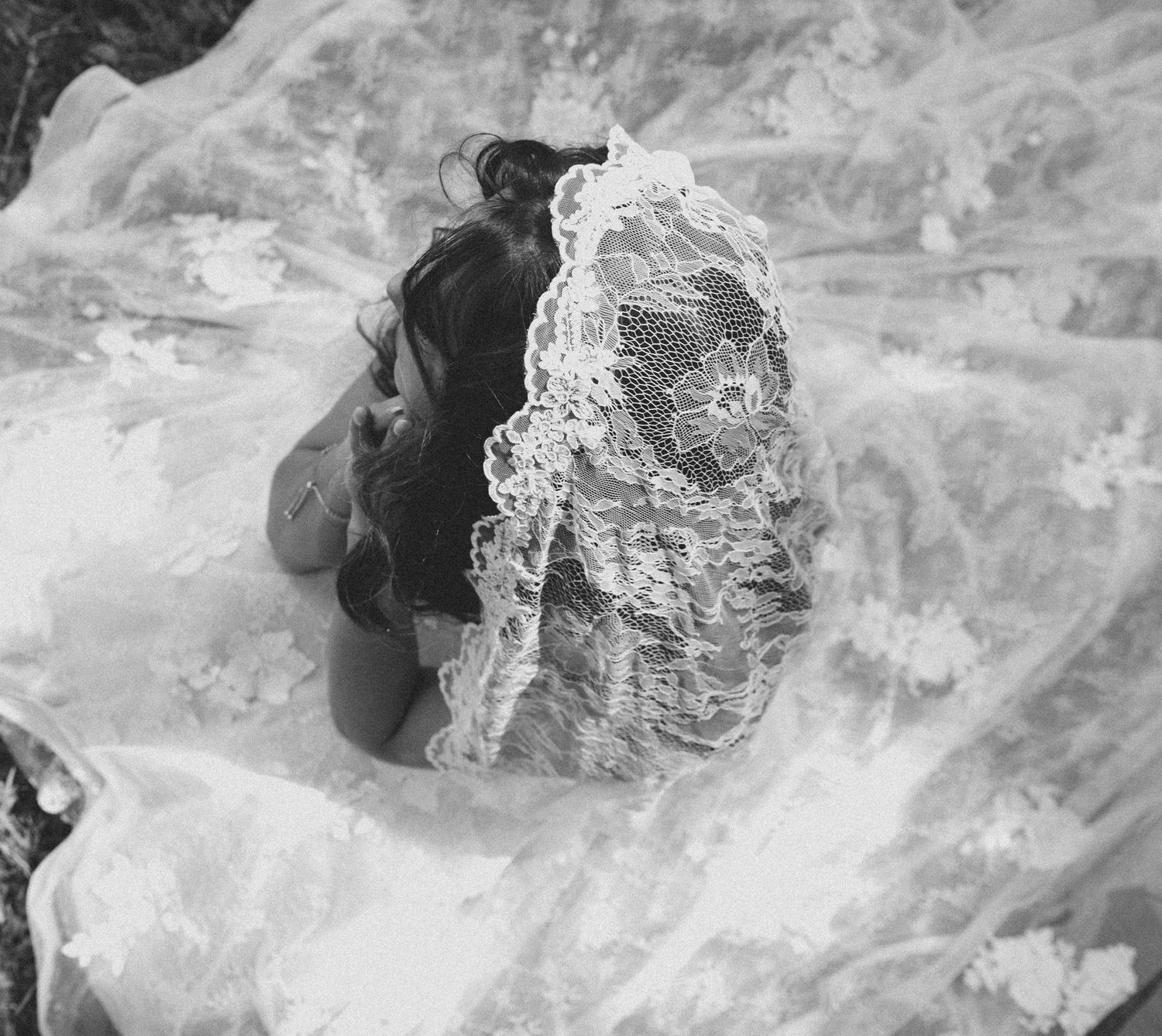 Black and white photo of a bride kneeling beneath a flowing lace wedding veil outdoors.