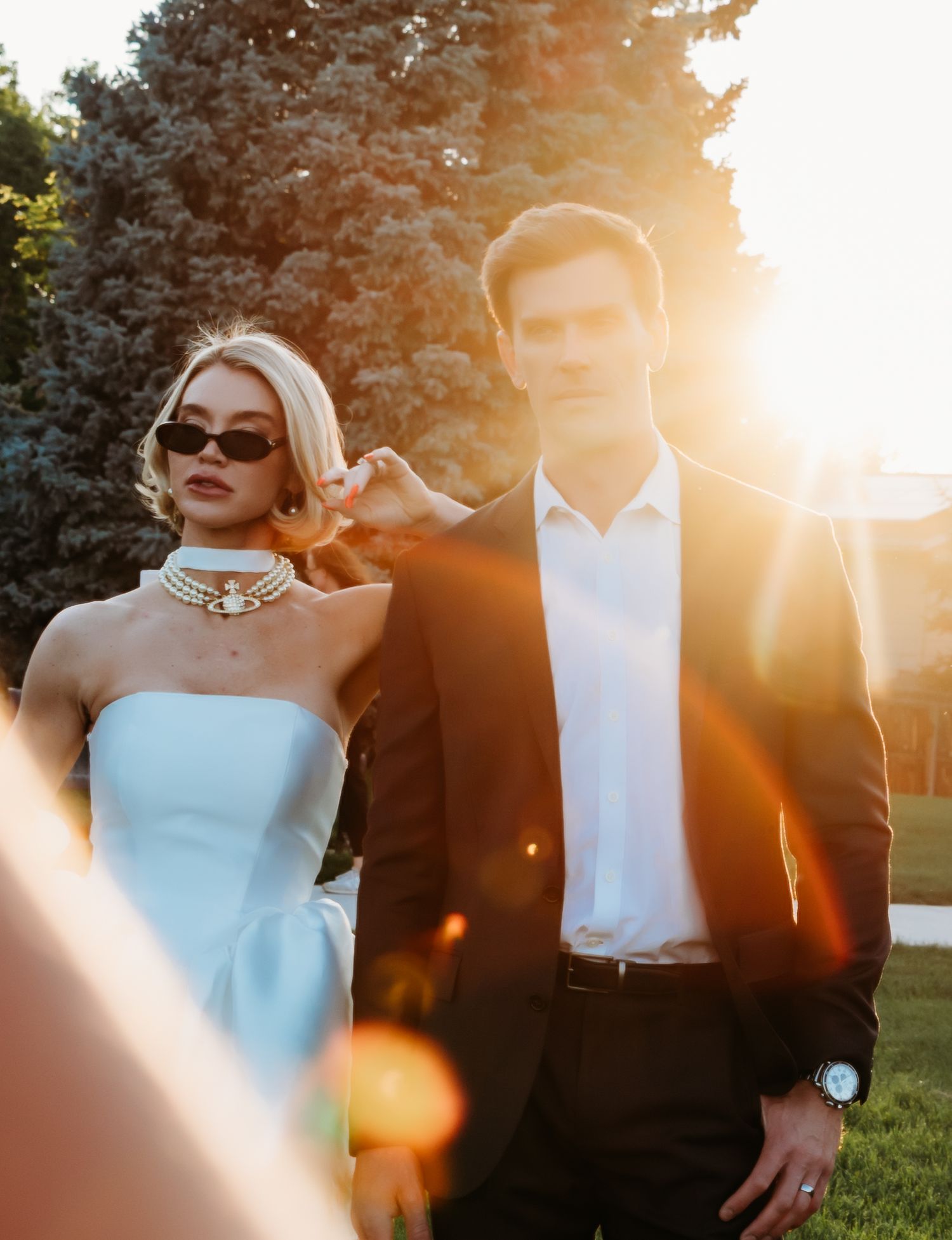 Golden sunset backlights couple in wedding attire creating a romantic silhouette against trees in garden setting.