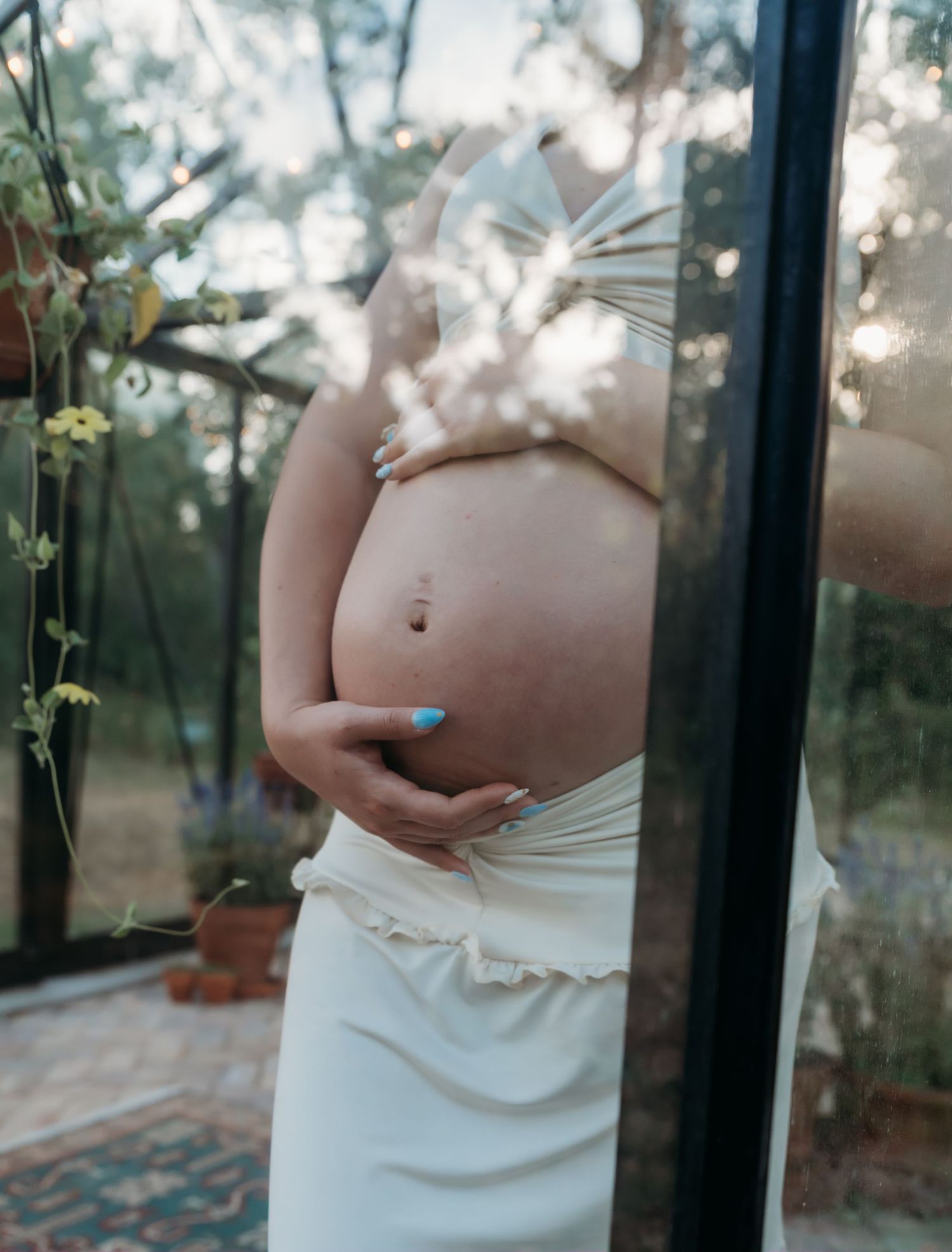 Maternity photo series showing pregnant belly against window with cherry blossoms and natural light creating dreamy atmosphere.