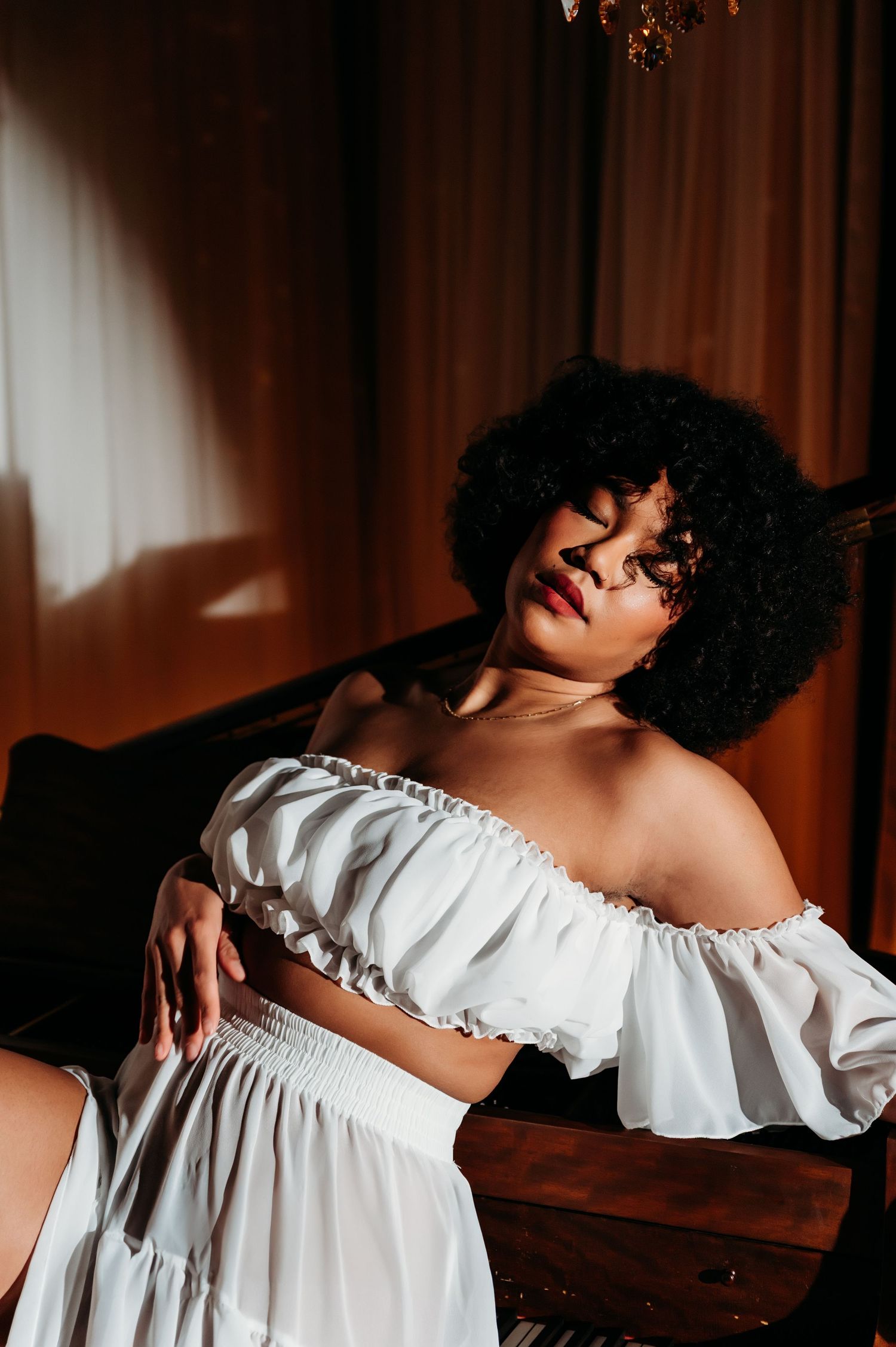 Dramatic portrait of a person in a white off-shoulder dress against warm lighting and curtains.