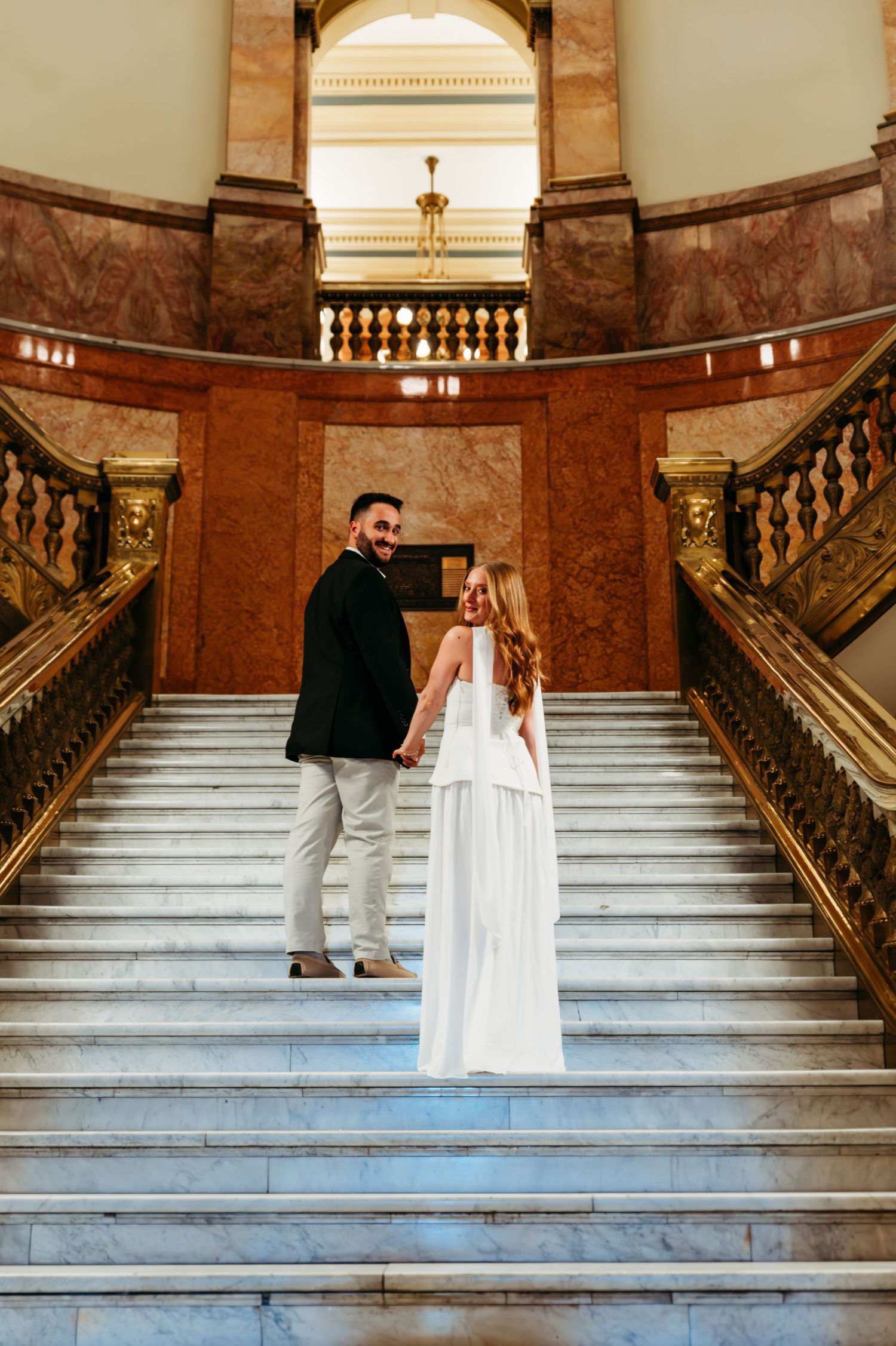 Couple in wedding attire holding hands on grand marble staircase in elegant venue with ornate gold railings.