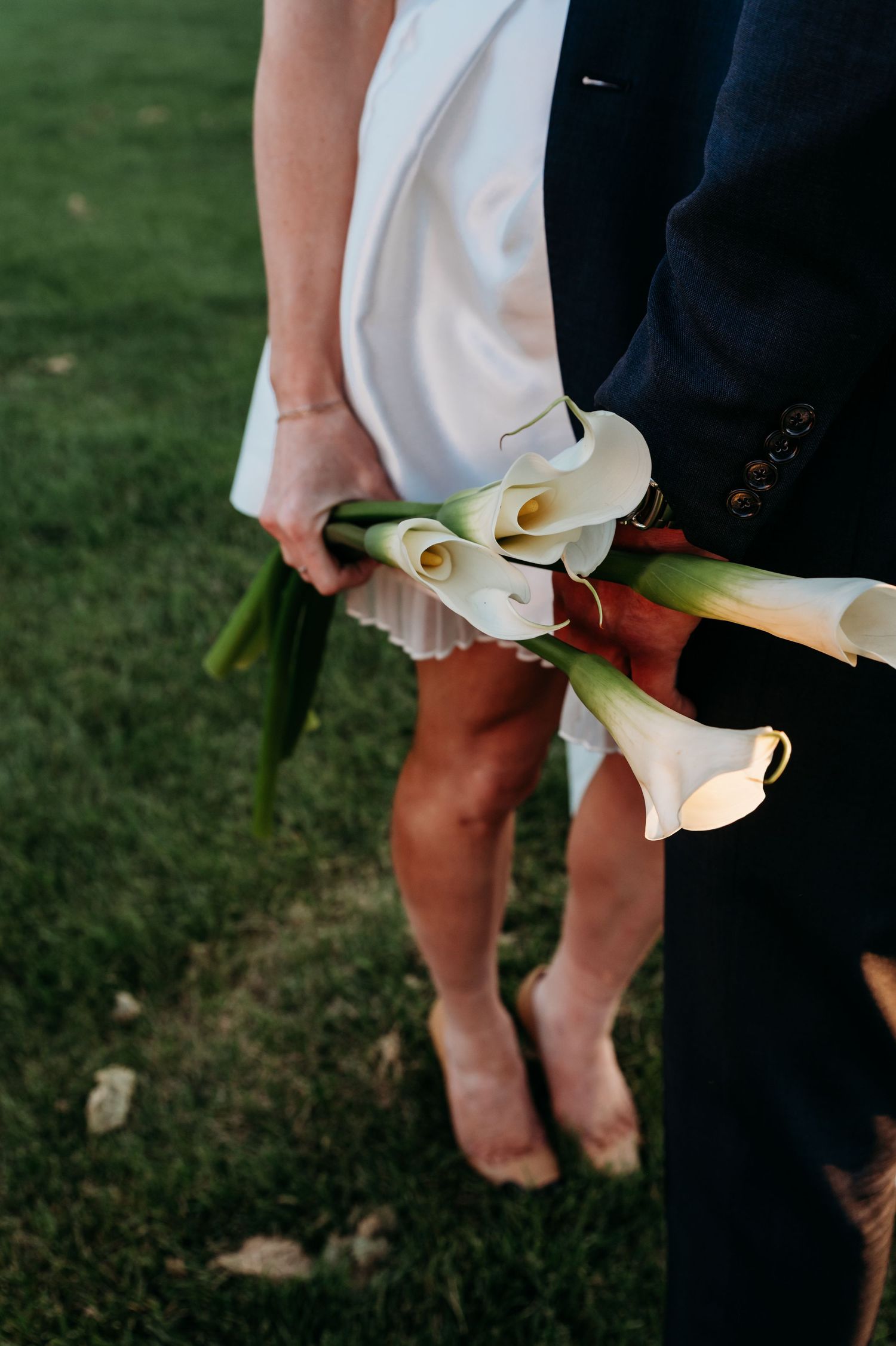 A white calla lily bouquet held against a dark suit and white dress on a grassy lawn during a wedding ceremony.