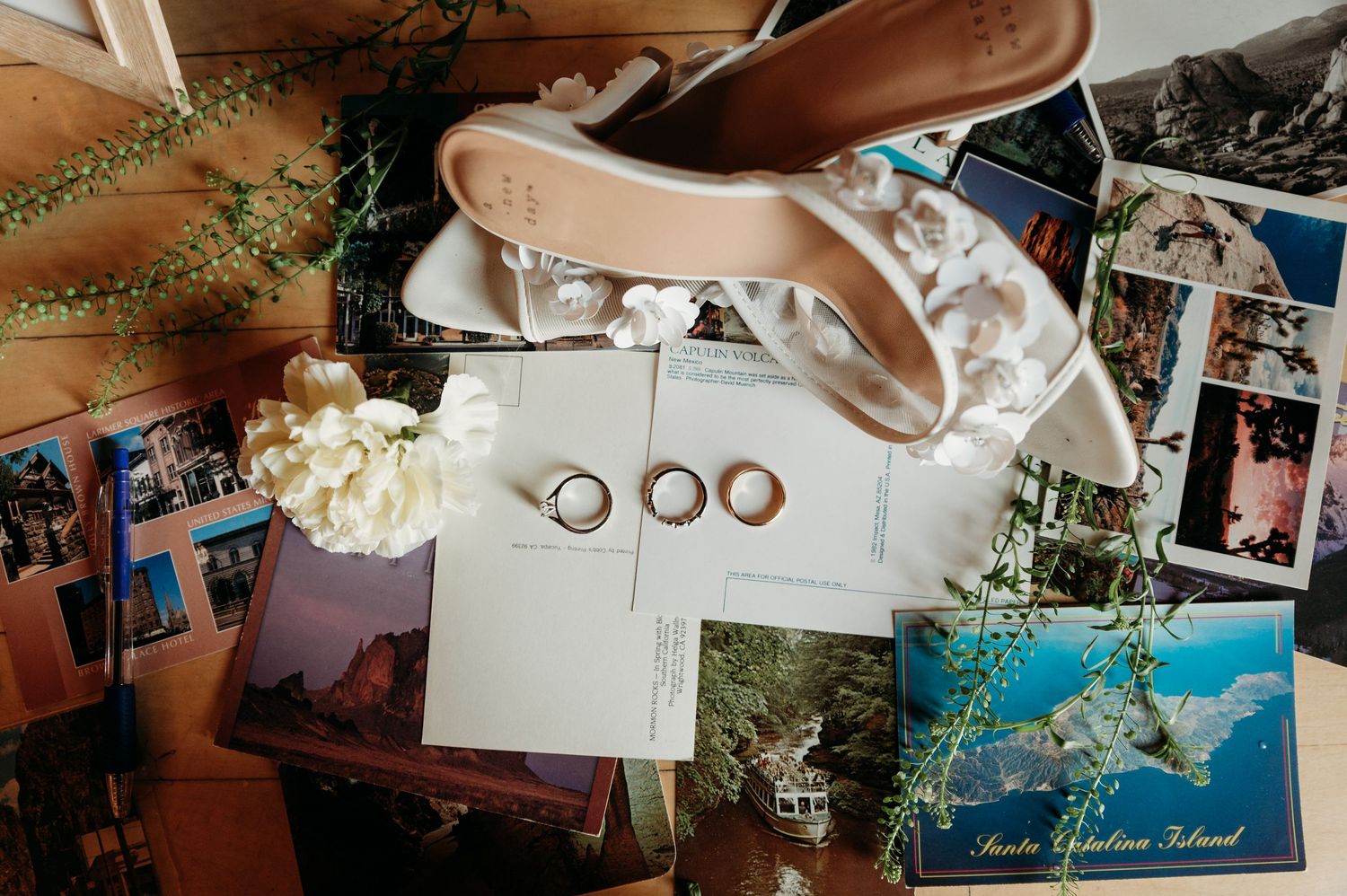 Wedding details flatlay with bridal shoes, rings, postcards and photographs scattered on wooden surface.