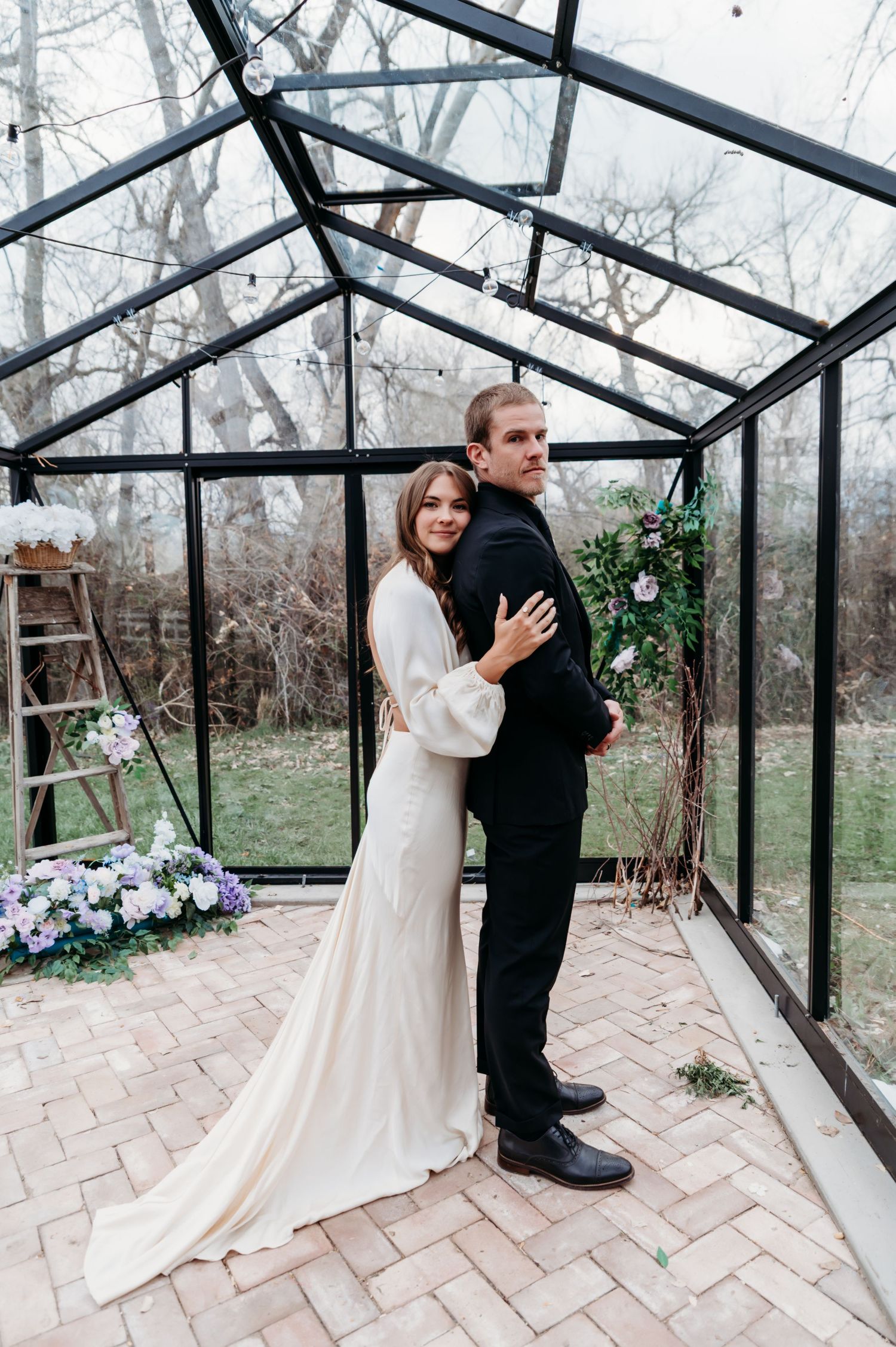 A couple shares intimate moments in a greenhouse setting with glass walls and brick flooring.