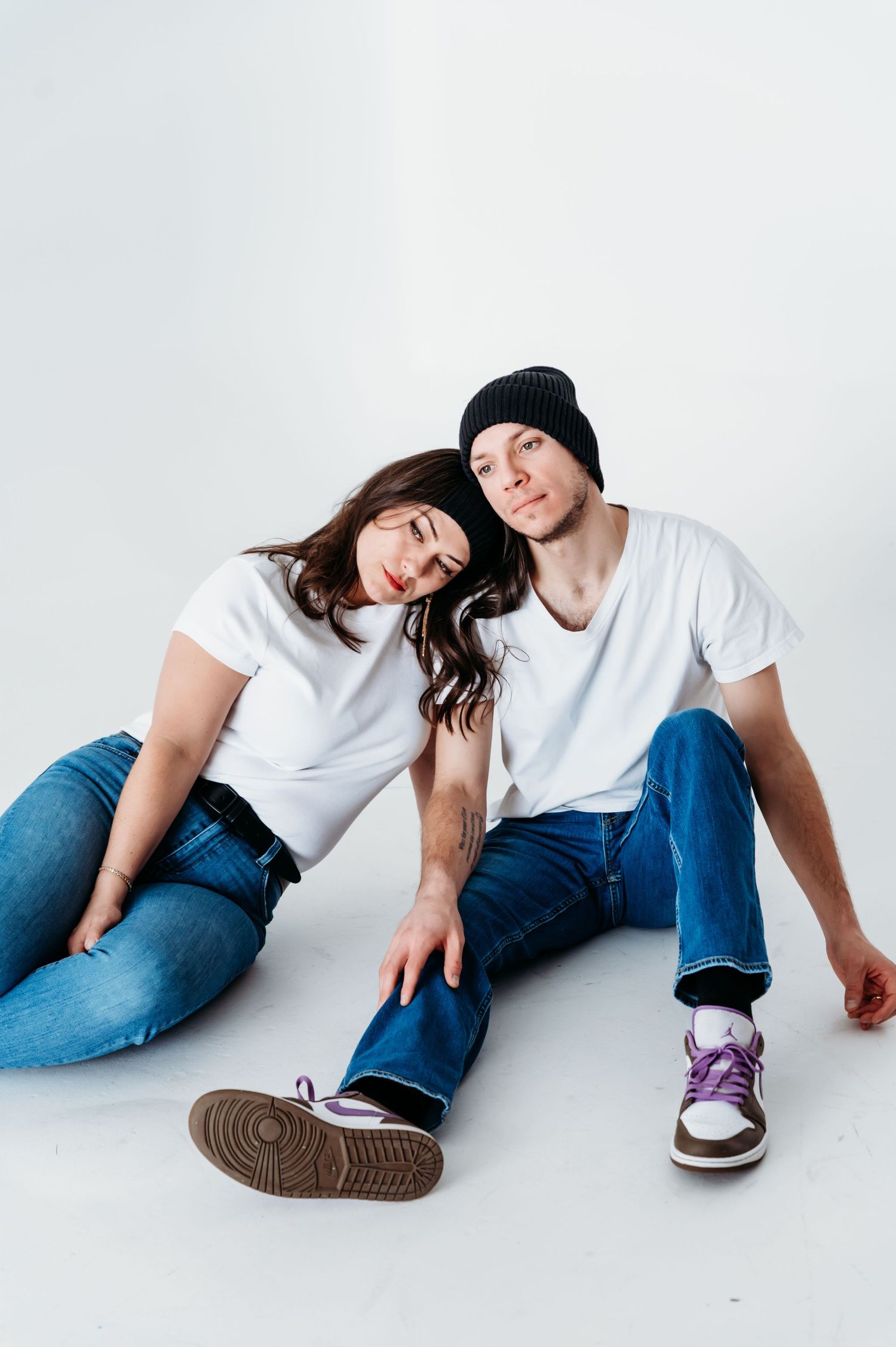 Two people in casual white t-shirts and jeans sit together on a white floor, wearing sneakers and a beanie.