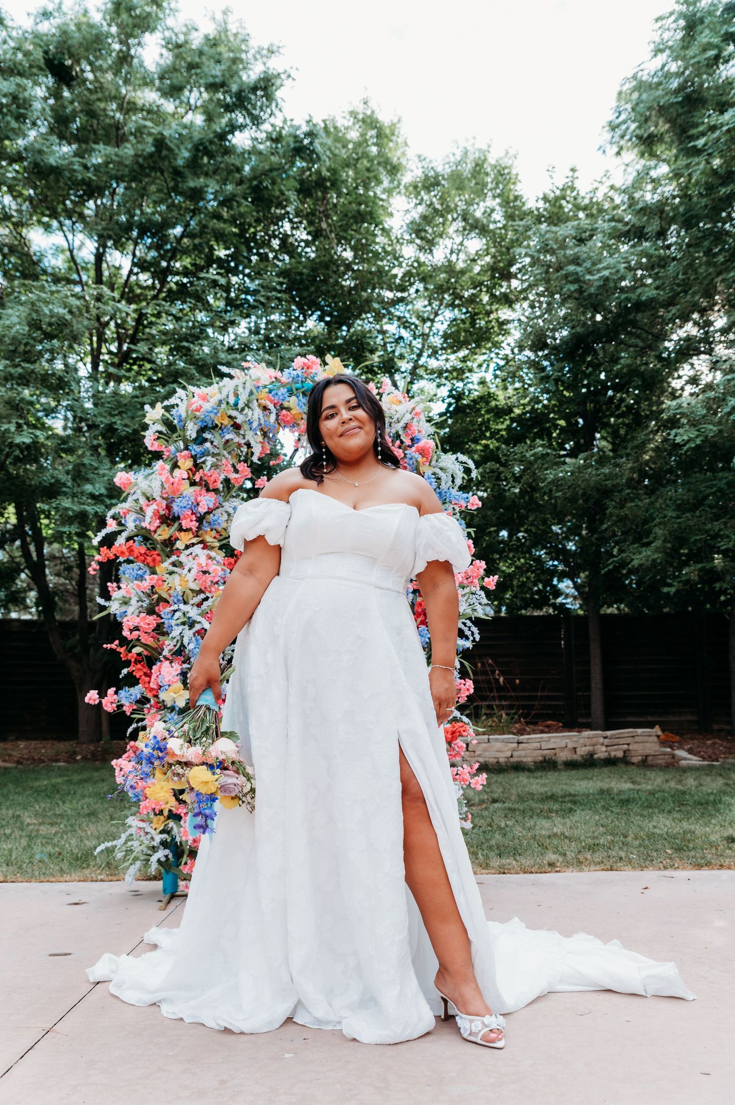 A bride in a white off-shoulder lace wedding dress poses in front of a floral arch with pink and blue blooms outdoors.