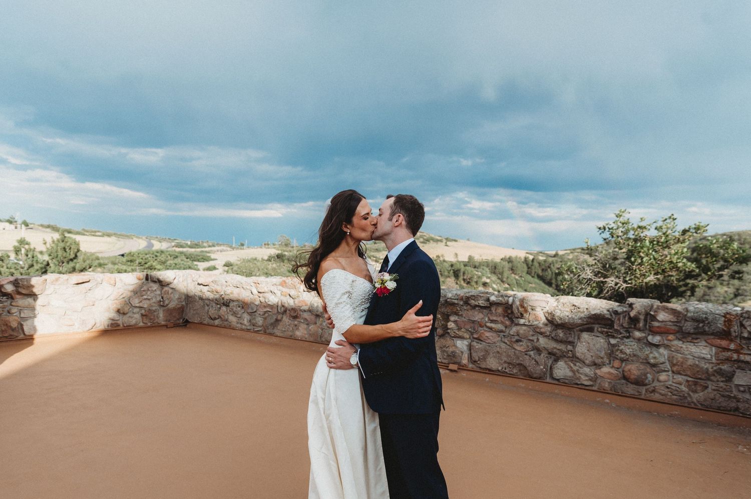 A bride and groom share a romantic kiss at an outdoor wedding venue with a stunning desert landscape and dramatic cloudy sky.