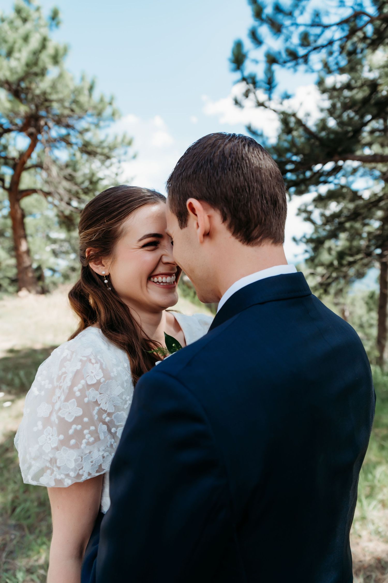 A joyful moment between a bride in lace and groom among pine trees.